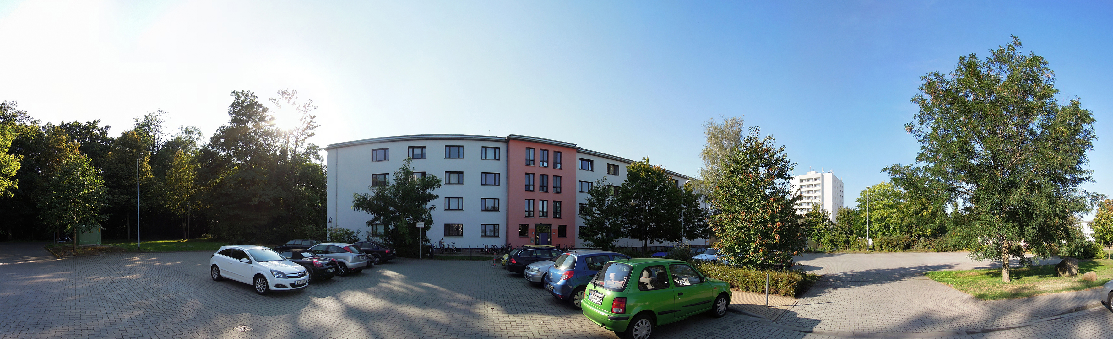 Ein mehrstöckiges Wohngebäude mit mehreren geparkten Autos davor. Das Gebäude hat eine weiße und eine pinkfarbene Fassade. Im Hintergrund sind Bäume und weitere Gebäude zu sehen. Der Himmel ist klar und blau.
