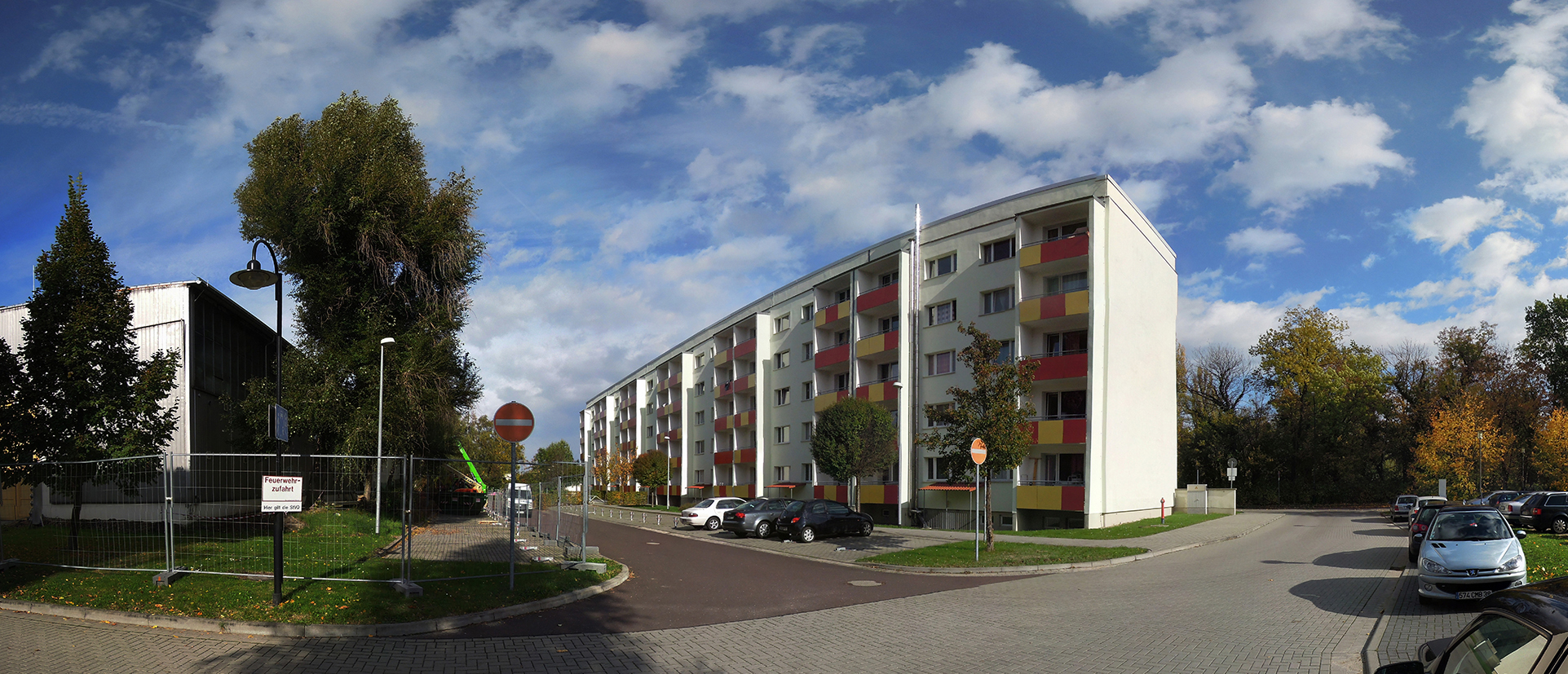 Ein Wohngebiet mit mehreren mehrstöckigen Wohnhäusern, geparkten Autos und Bäumen. Die Straße ist mit Kopfsteinpflaster belegt und es gibt Verkehrsschilder. Der Himmel ist teilweise bewölkt.