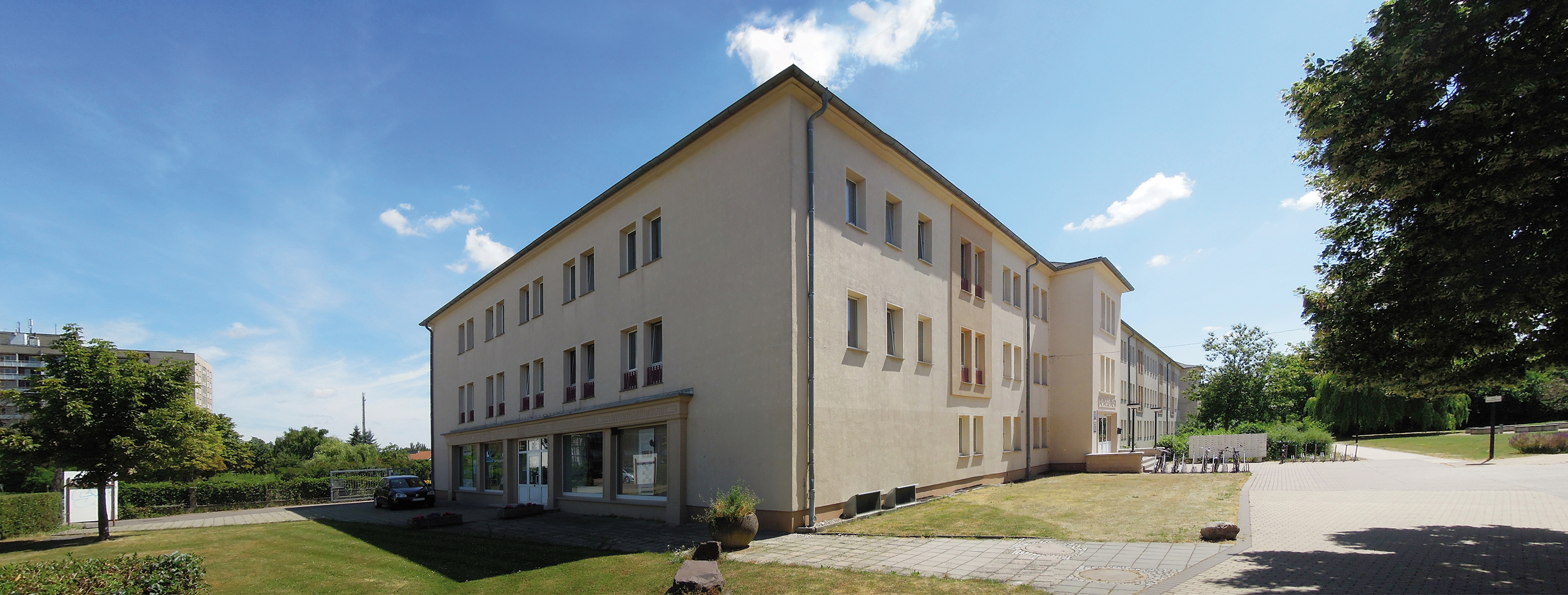 Ein mehrstöckiges Gebäude mit hellen Wänden und vielen Fenstern steht auf einem gepflegten Gelände. Vor dem Gebäude befindet sich ein gepflasterter Bereich mit einigen Tischen und Stühlen. Im Hintergrund sind Bäume und weitere Gebäude zu sehen. Der Himmel ist blau mit einigen Wolken.