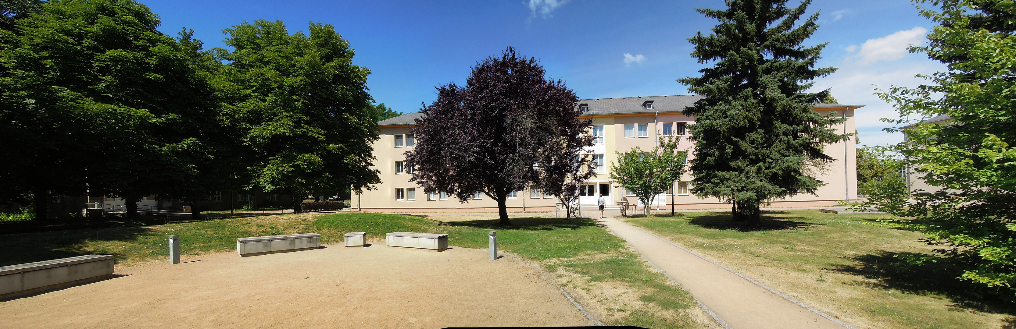 Ein Park mit einem Sandspielplatz, Bänken und Wegen. Im Hintergrund steht ein mehrstöckiges Gebäude, umgeben von Bäumen und Grünflächen. Der Himmel ist blau mit einigen Wolken.