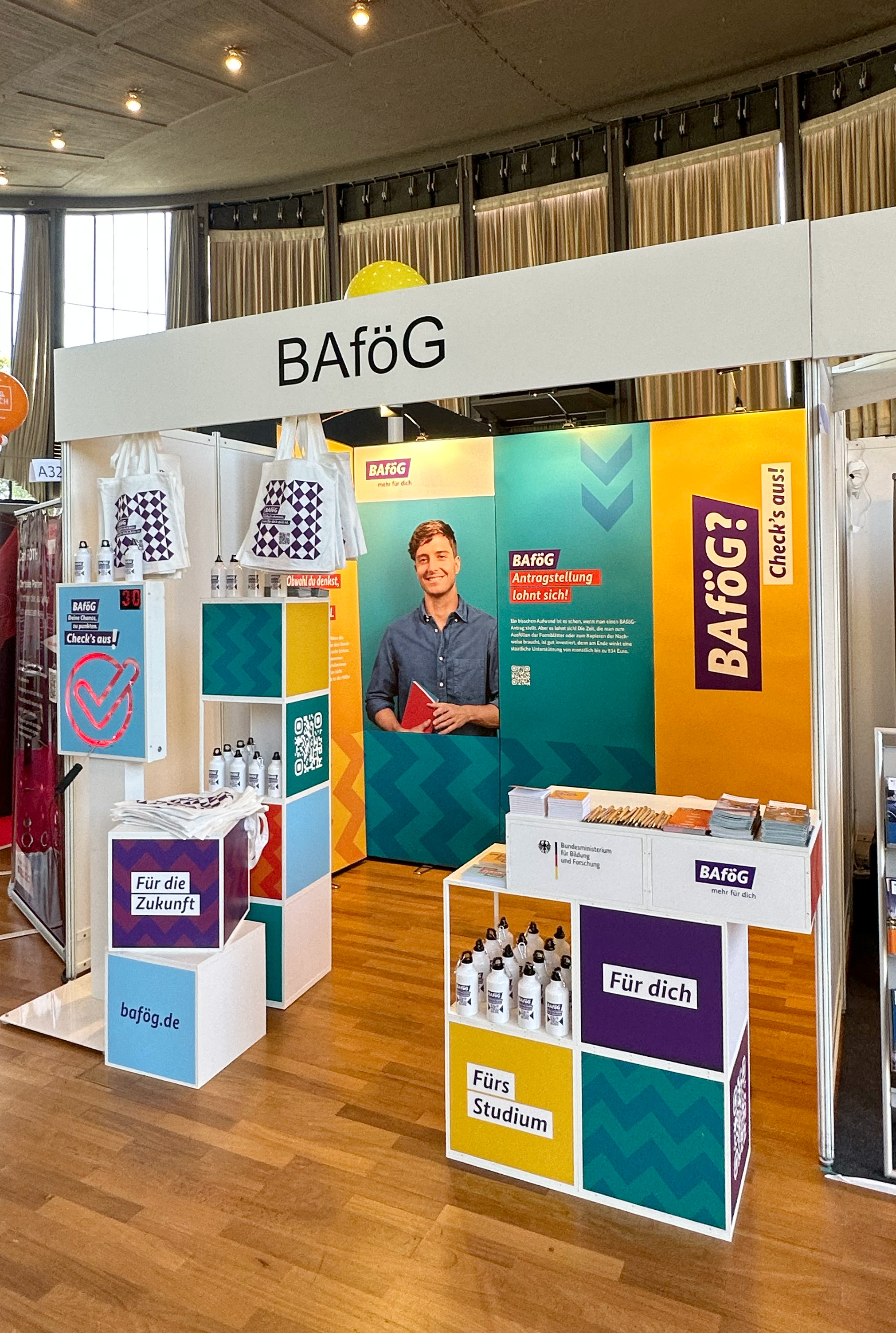 Ein BAföG-Informationsstand mit Broschüren, Postern und einem Plakat mit einem lächelnden jungen Mann. Der Stand bietet Informationen über BAföG-Förderung für Studierende, mit dem Slogan "Einfach für dich" und dem Hinweis "Check's aus!". Die Website "www.bafög.de" ist sichtbar.
