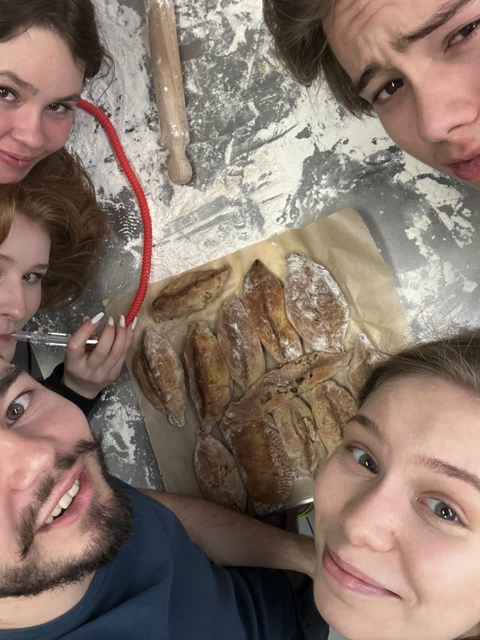 Eine Gruppe von vier Personen lächelt in die Kamera, während sie gemeinsam ein großes Stück Brot oder Kuchen teilt, das in Alufolie eingewickelt ist. Sie wirken fröhlich und genießen den Moment.
