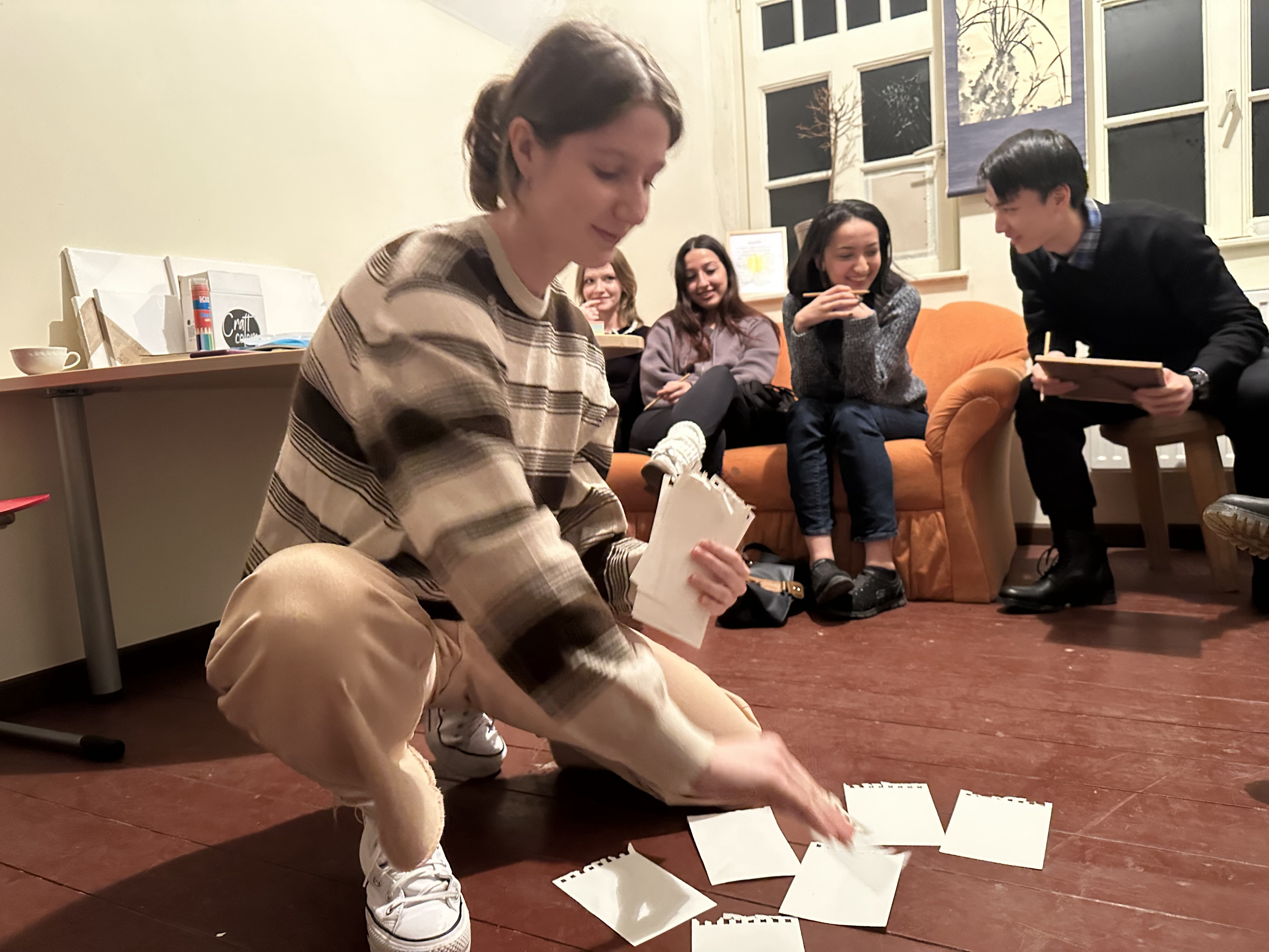 Eine Frau kniet auf dem Boden und sortiert mehrere Zettel. Im Hintergrund sitzen drei Personen auf einem Sofa und beobachten sie. Die Szene scheint in einem Raum mit Büchern und einem Tisch zu sein.