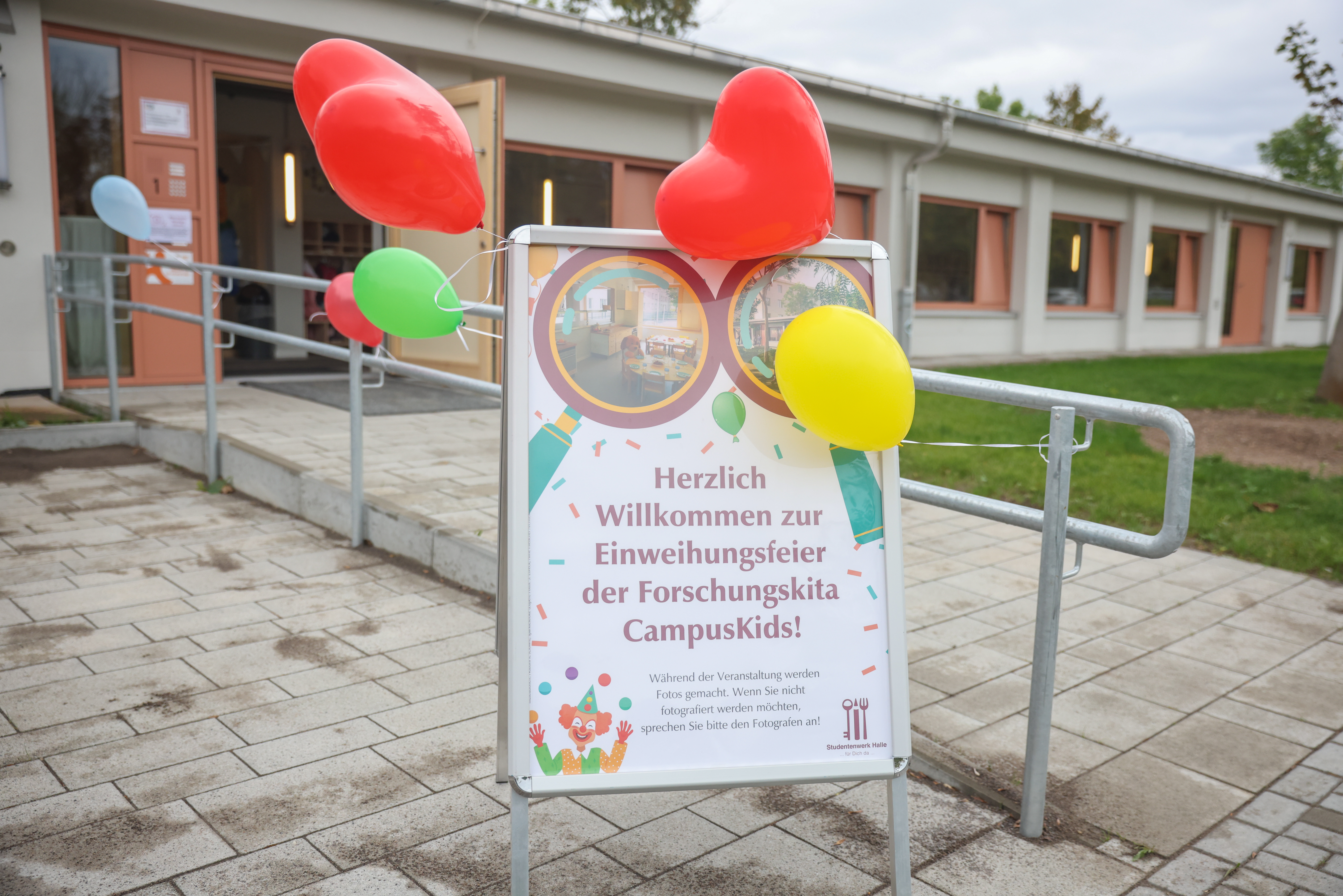 "Einladungsschilder mit Luftballons vor einem Gebäude. Das Schild begrüßt zur Einweihungsfeier des Forschungskitas CampusKids. Es steht auf einem Gehweg mit einer Rampe im Hintergrund. Herzlich willkommen zur Einweihungsfeier der Forschungskita CampusKids!"