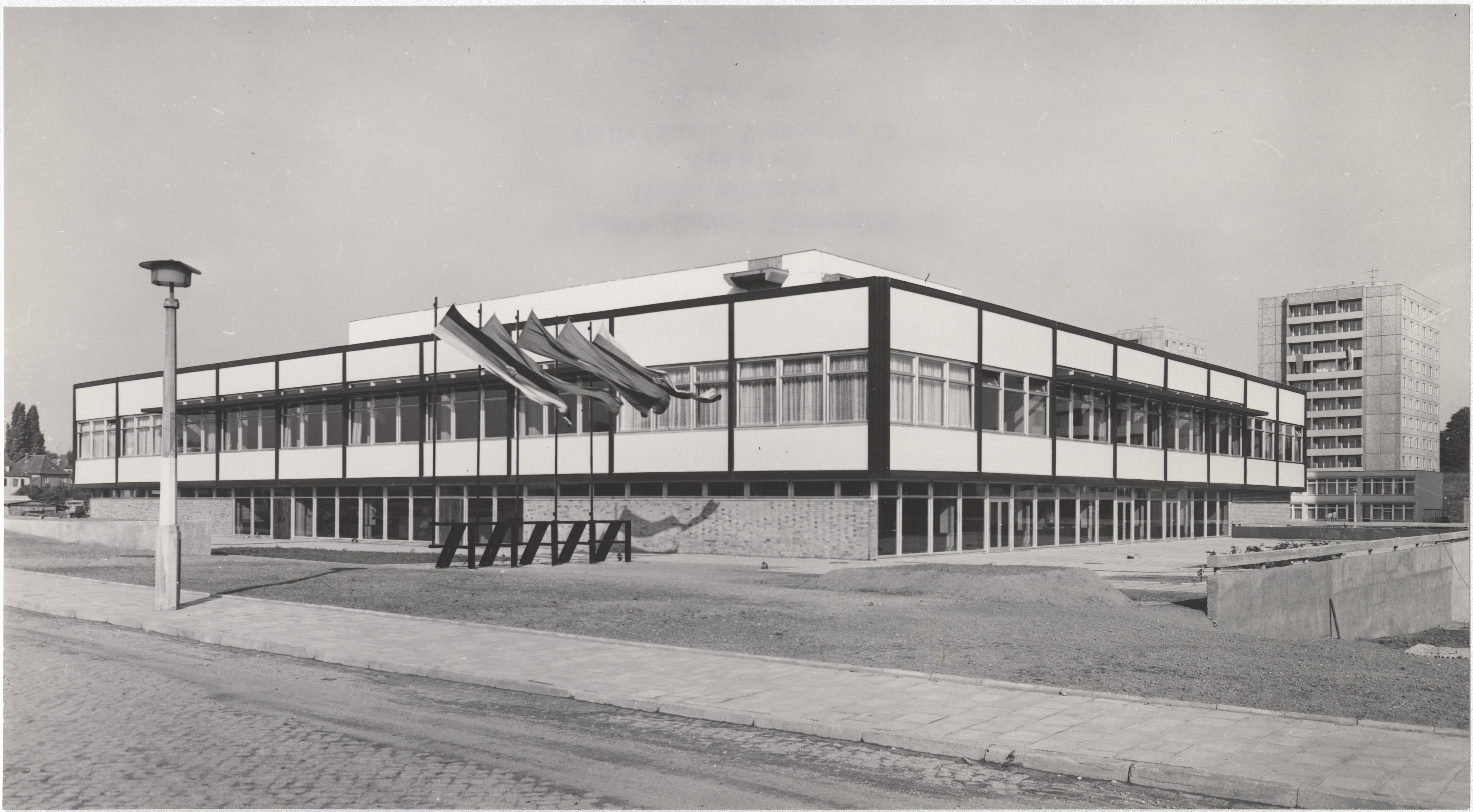 Ein modernes Gebäude mit großen Fenstern und einer markanten Dachkonstruktion. Im Vordergrund befindet sich ein Gehweg mit einer Straßenlaterne. Im Hintergrund sind weitere Gebäude zu sehen. Das Foto ist in Schwarz-Weiß gehalten.