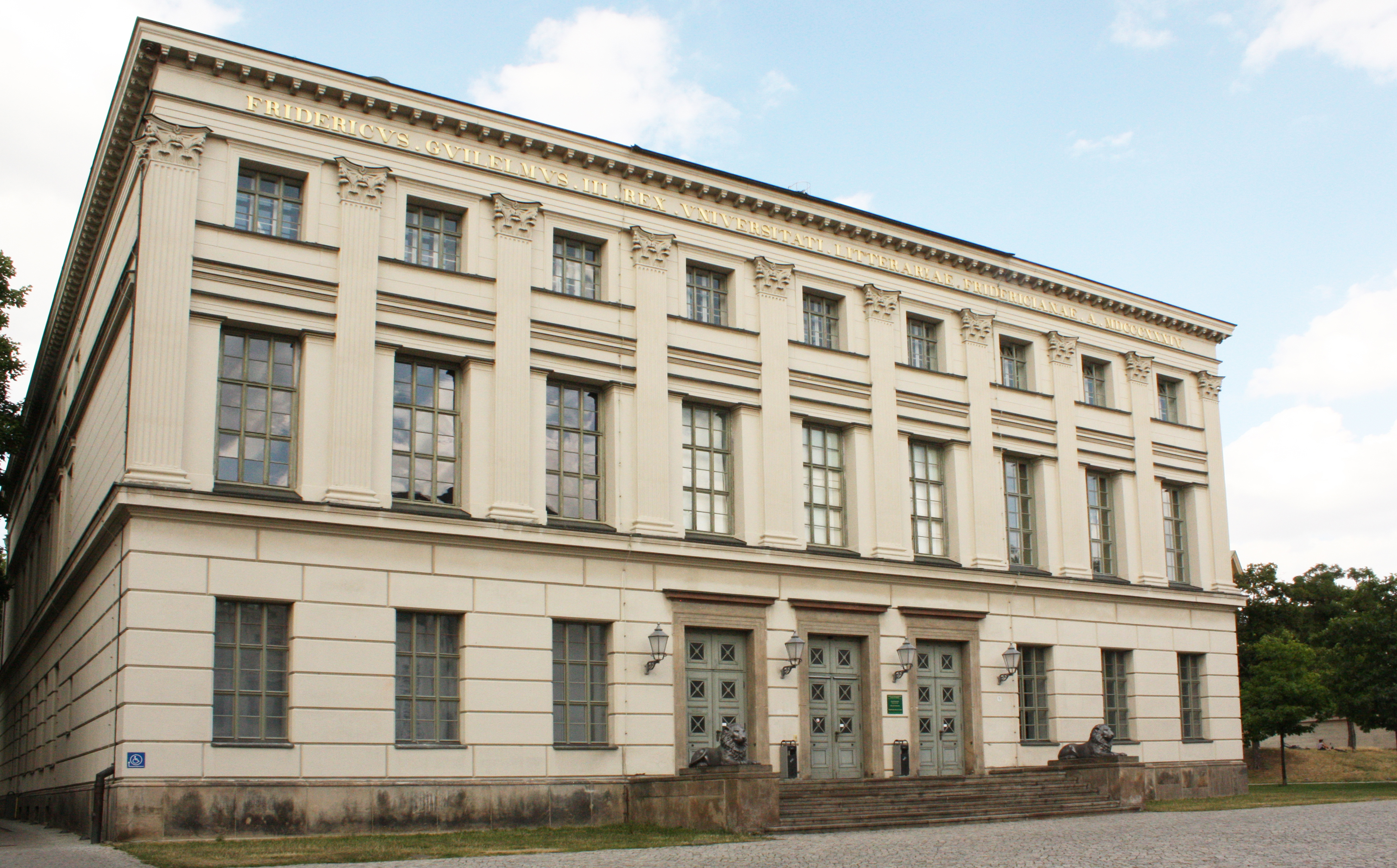 Ein großes, klassizistisches Gebäude mit hellbeiger Fassade und vielen großen Fenstern. Das Gebäude hat zwei Stockwerke und einen symmetrischen Aufbau. Vor dem Gebäude befindet sich ein kleiner Vorplatz mit einem gepflasterten Bereich.