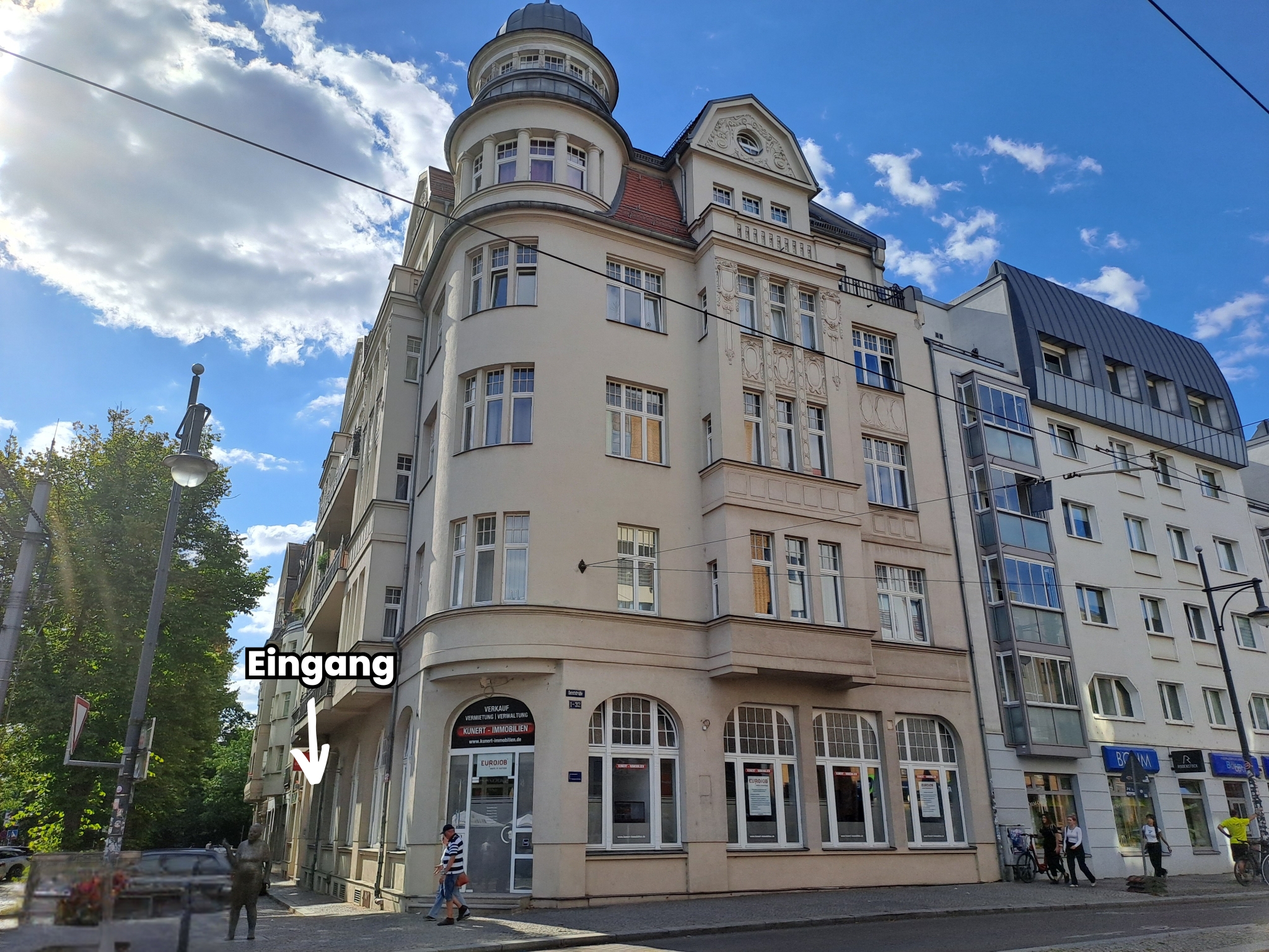 "Ein mehrstöckiges Gebäude mit markanter Architektur, das an einer Straßenecke steht. Der Eingang ist mit einem Schild gekennzeichnet. Das Gebäude hat große Fenster und eine runde Ecke. Im Hintergrund sind weitere moderne Gebäude und ein klarer Himmel mit einigen Wolken zu sehen."