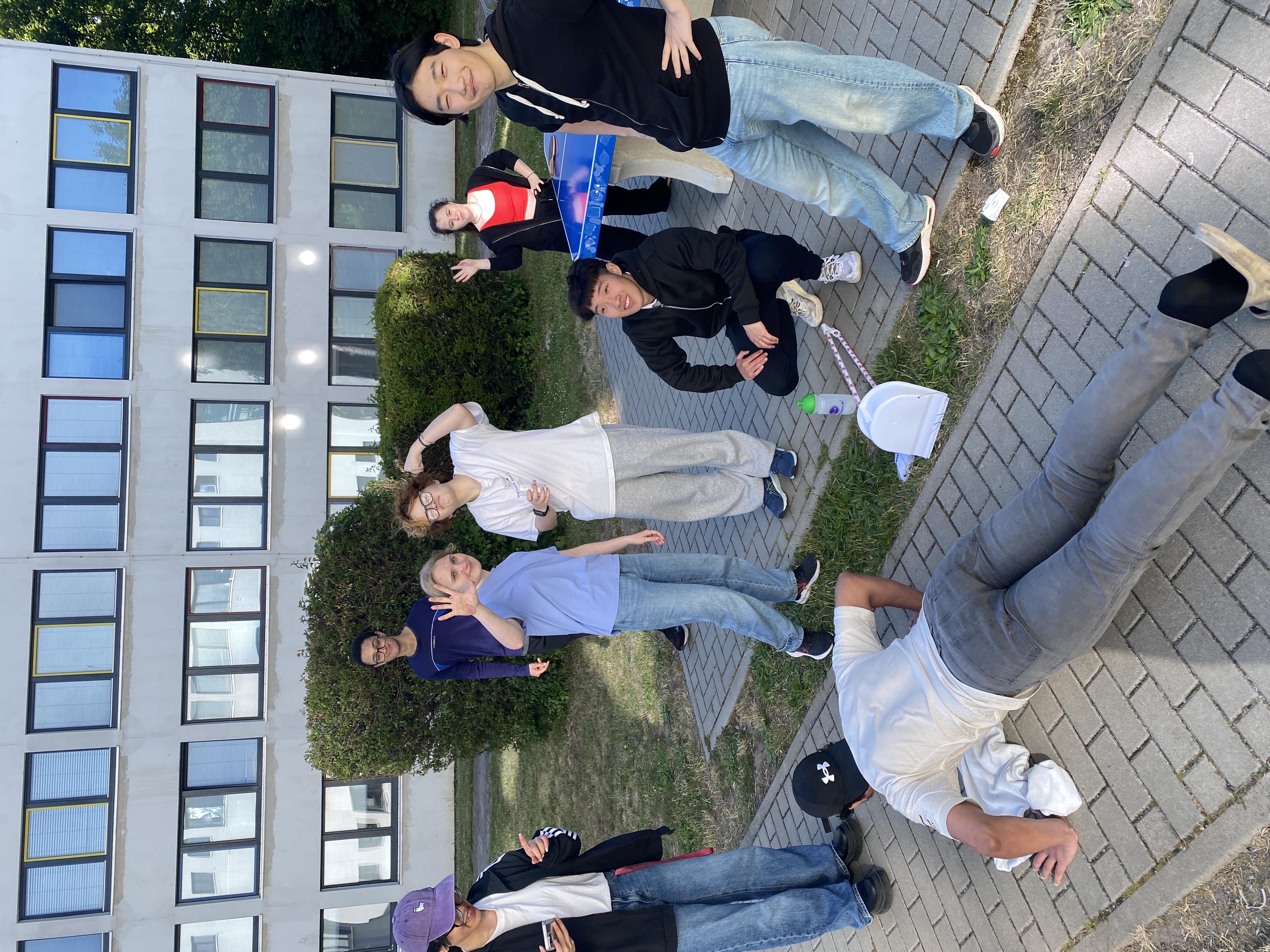 Eine Gruppe junger Menschen steht und sitzt vor einem Gebäude. Einige posieren für das Foto, während eine Person auf dem Boden liegt. Sie wirken entspannt und fröhlich, einige machen Handzeichen oder lächeln. Die Szene wirkt wie ein lockeres Treffen oder eine Pause.