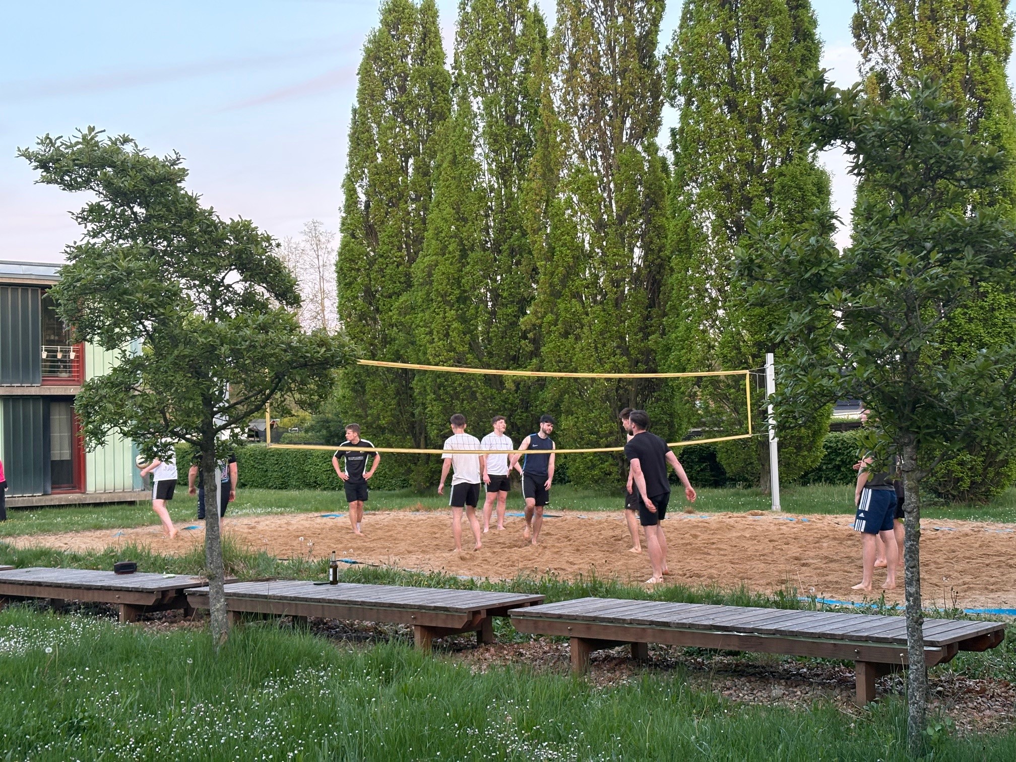 "Eine Gruppe von Menschen spielt Volleyball auf einem Sandplatz im Freien. Im Hintergrund sind Bäume und ein Gebäude zu sehen. Die Spieler sind in Bewegung und scheinen Spaß zu haben."