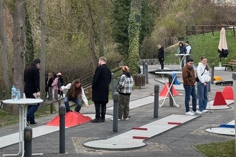 "Menschen spielen auf einem Mini-Golf-Platz im Freien. Der Platz ist mit Hindernissen und Kurven ausgestattet. Die Spieler sind auf einem gepflasterten Weg verteilt und konzentrieren sich auf das Spiel. Im Hintergrund sind Bäume und eine Wiese zu sehen."
