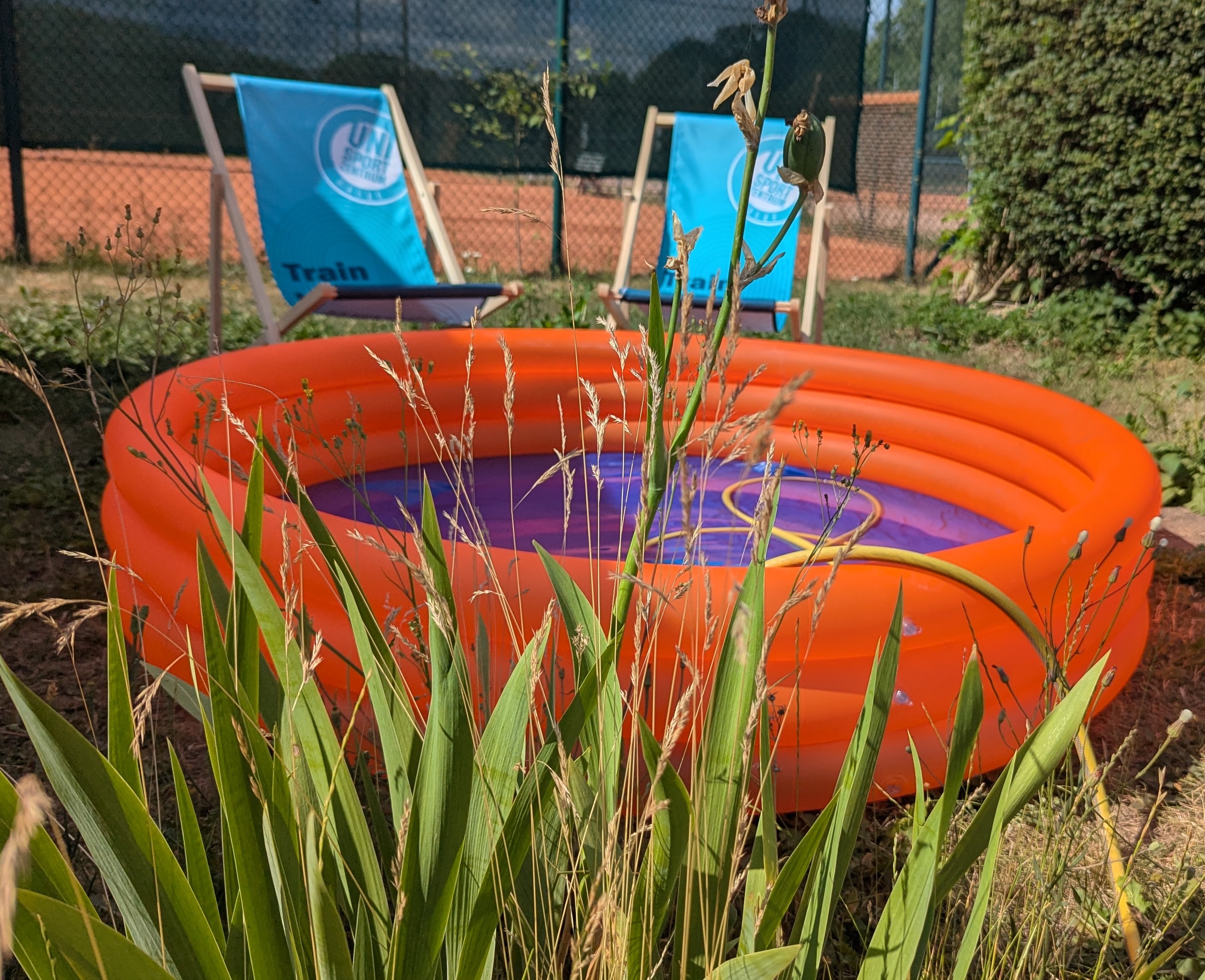 Ein orangefarbener aufblasbarer Pool mit einem blauen Design im Inneren steht auf einem Rasen. Davor sind zwei blaue Liegestühle mit Werbung für "Train" aufgestellt. Der Pool ist von hohem Gras umgeben und befindet sich in einem Gartenbereich.