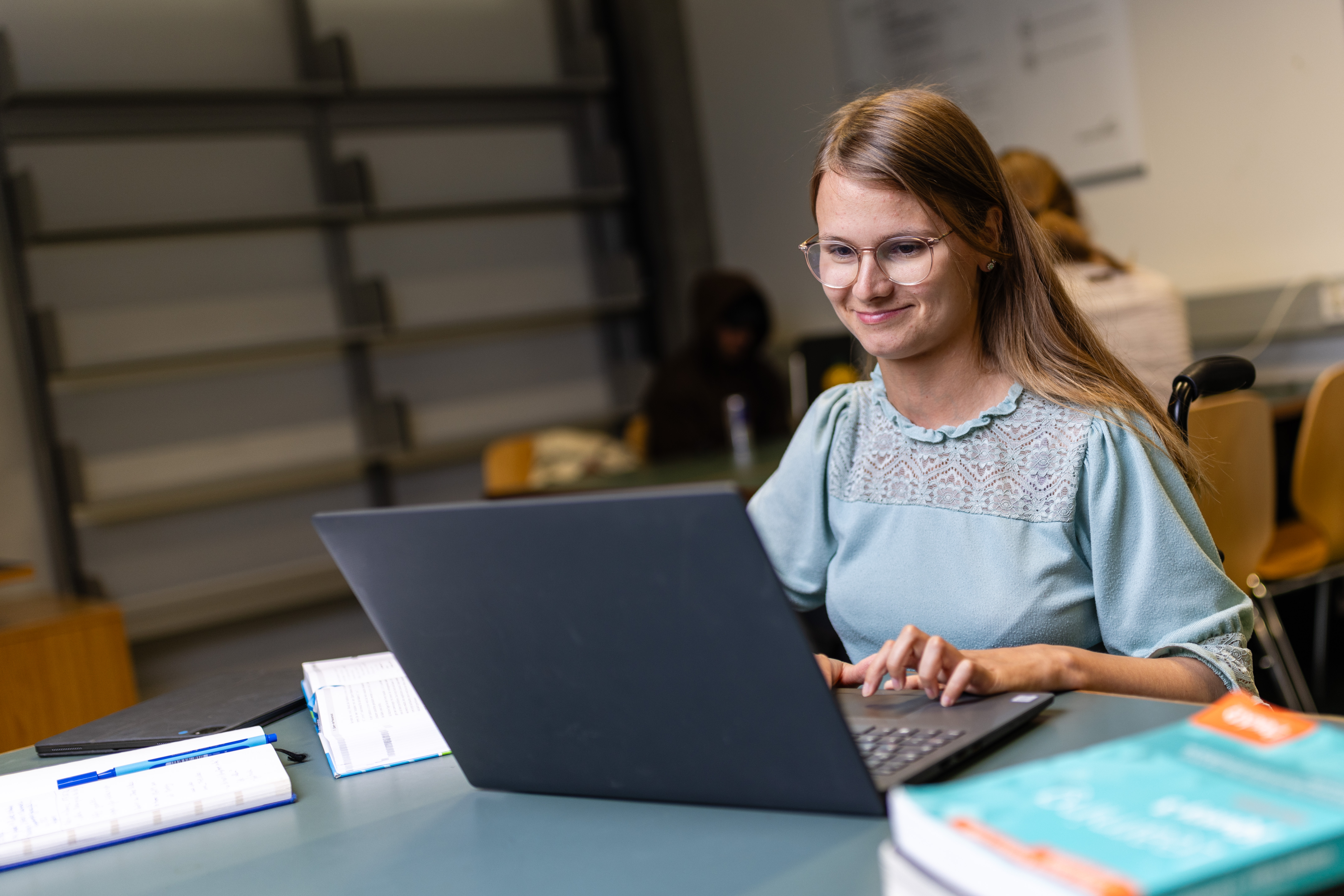 Eine Frau mit Brille sitzt an einem Tisch und arbeitet konzentriert an einem Laptop. Vor ihr liegen Bücher und Notizen. Sie trägt ein helles Oberteil und lächelt leicht. Der Raum wirkt wie ein ruhiger Arbeits- oder Studienbereich.