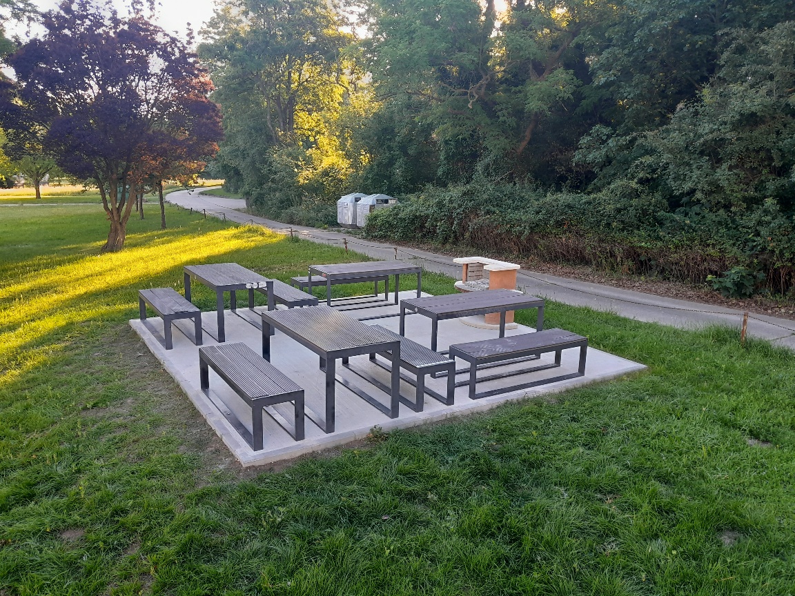Moderne Sitzanlage aus mehreren Betonplatten mit integrierten Bänken und Tischen in geometrischer Anordnung und gemauertem Grill auf Rasenfläche, umgeben von Bäumen und Grünflächen.