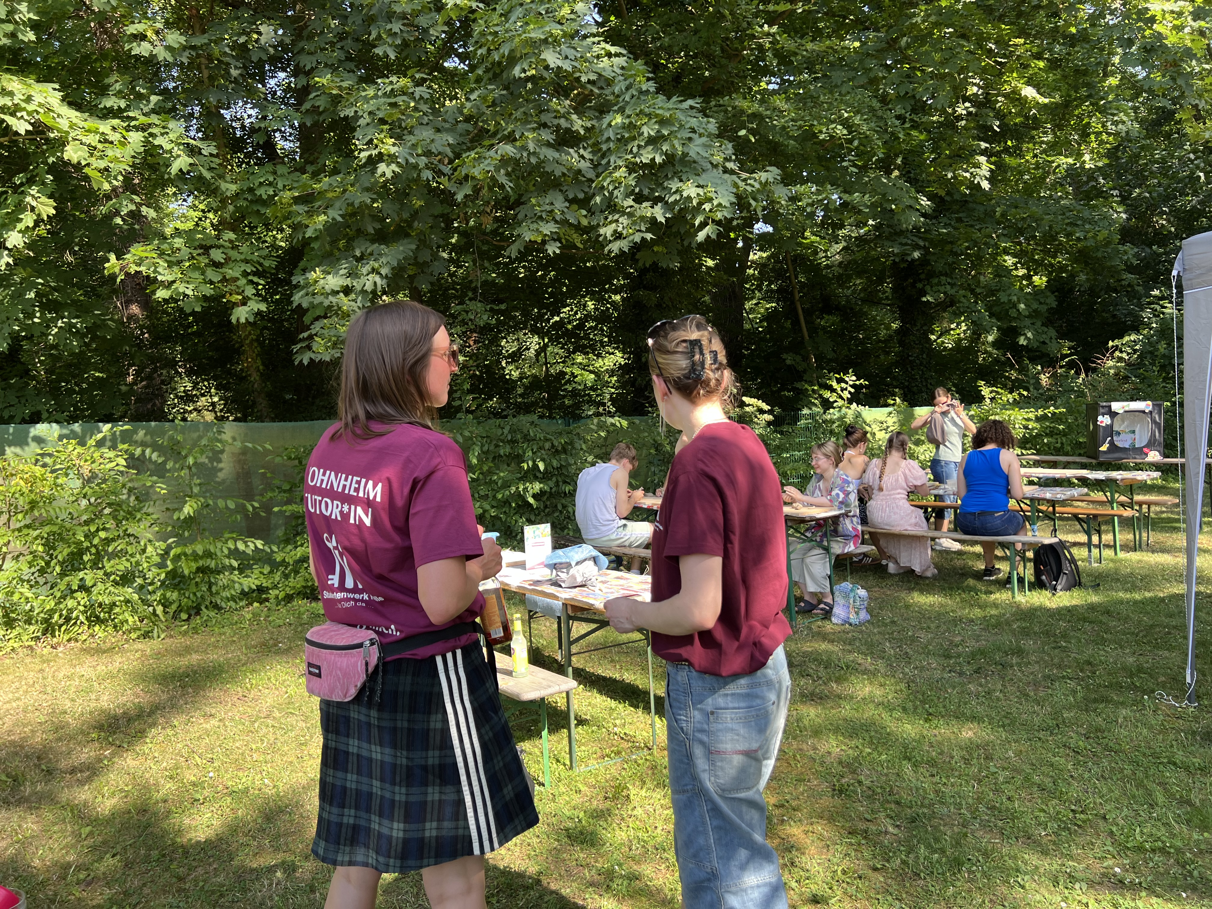 Zwei Personen stehen im Freien an einem Tisch mit Unterlagen und unterhalten sich. Die Person links trägt ein dunkelrotes T-Shirt mit Aufdruck und eine karierte Schürze, die Person rechts ein rotes Shirt und Jeans. Im Hintergrund sitzen weitere Menschen an Tischen unter Bäumen.