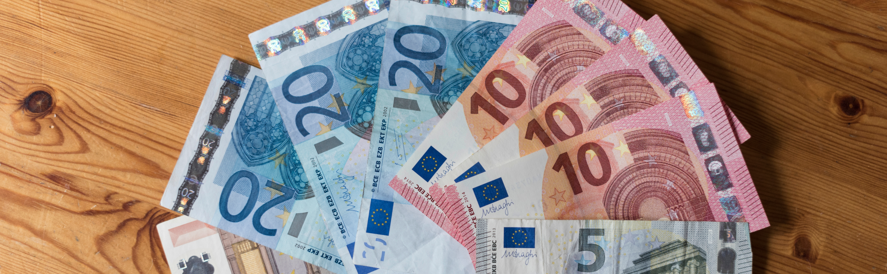 "Ein Stapel Euro-Banknoten verschiedener Denominationen liegt auf einem Holztisch. Die Scheine sind 5, 10, 20, 50 und 100 Euro. Die Banknoten sind teilweise überlappend angeordnet und zeigen die typischen Sicherheitsmerkmale und Designs der Euro-Banknoten."