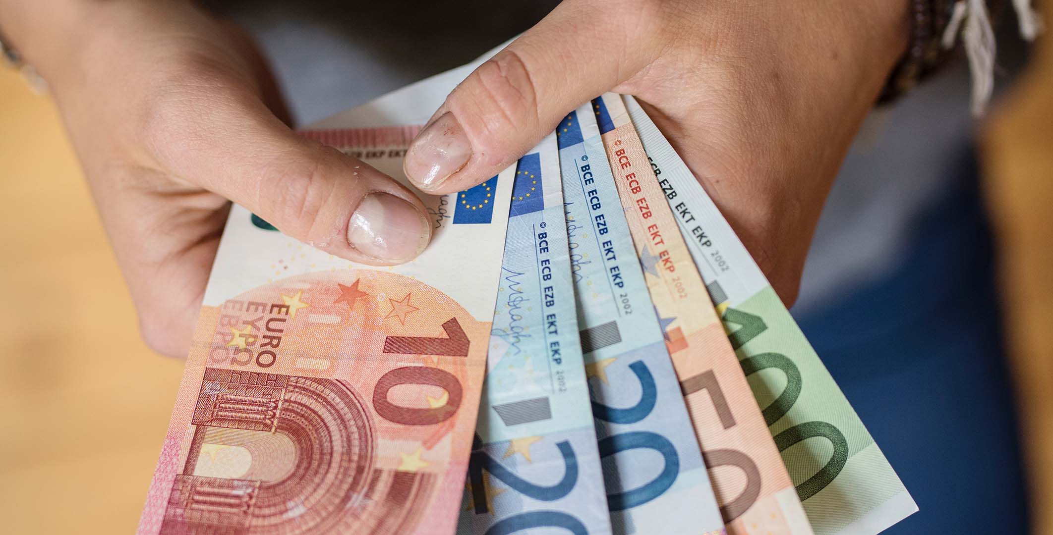 "Eine Hand hält verschiedene Euro-Scheine, darunter 10, 20, 50 und 100 Euro. Die Scheine sind fächerförmig angeordnet und zeigen die Vorderseiten mit den unterschiedlichen Designs und Farben. Die Hand hält die Scheine zwischen Daumen und Zeigefinger."