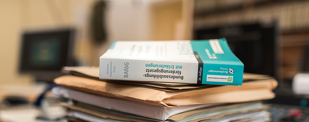 Ein Stapel Bücher und Papiere auf einem Tisch. Obenauf liegen zwei Bücher, eines mit weißem und eines mit blauem Einband. Die Bücher scheinen sich mit rechtlichen oder behördlichen Themen zu befassen, wie die Titel vermuten lassen.