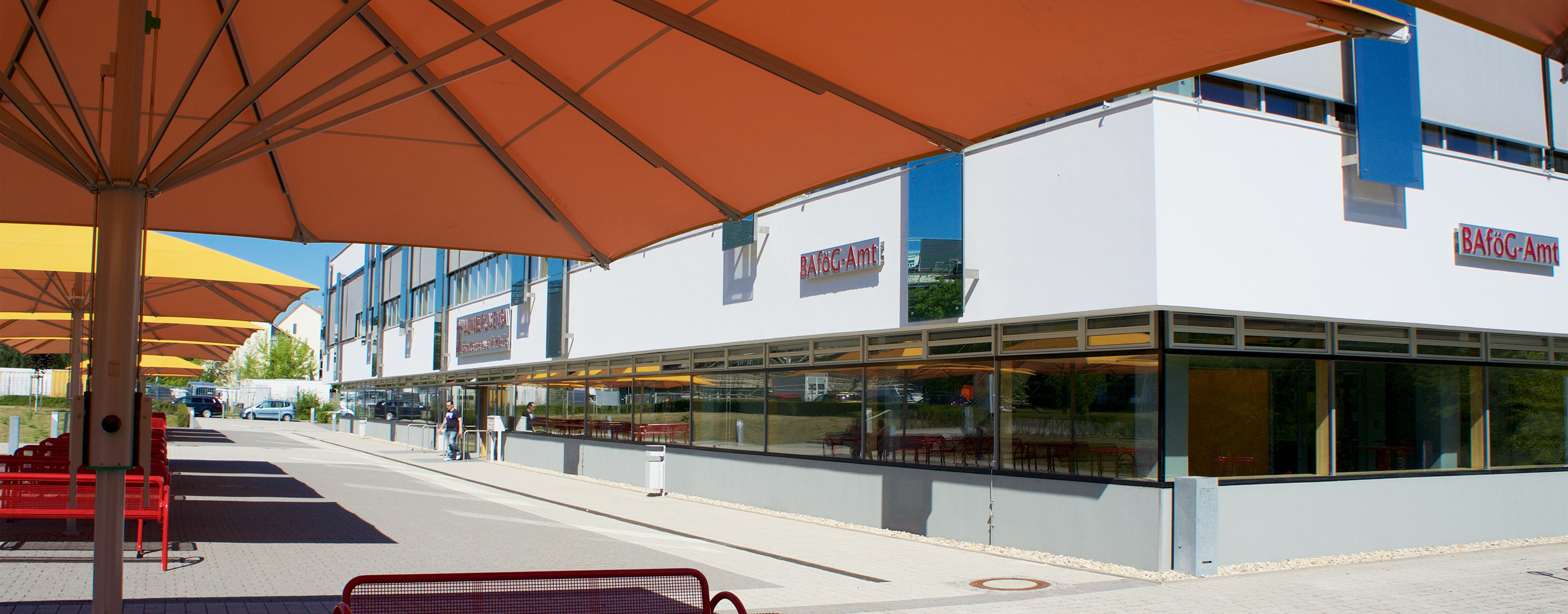 Ein modernes Einkaufszentrum mit mehreren Geschäften, darunter ein "Bauknecht"-Geschäft. Vor dem Gebäude befinden sich große Sonnenschirme und eine breite, leere Straße. Im Hintergrund sind weitere Geschäfte und ein paar Menschen zu sehen.