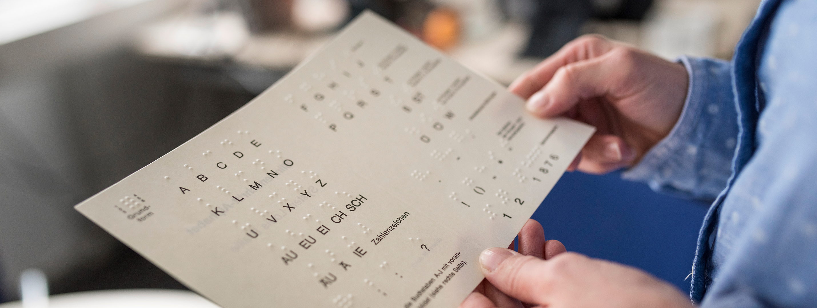 Eine Person hält ein Blatt mit dem Brailleschriftsystem in den Händen. Das Blatt zeigt die Braille-Zeichen für den Alphabetbuchstaben A bis Z, Zahlen von 0 bis 9 sowie einige Sonderzeichen und Anweisungen zur Benutzung.