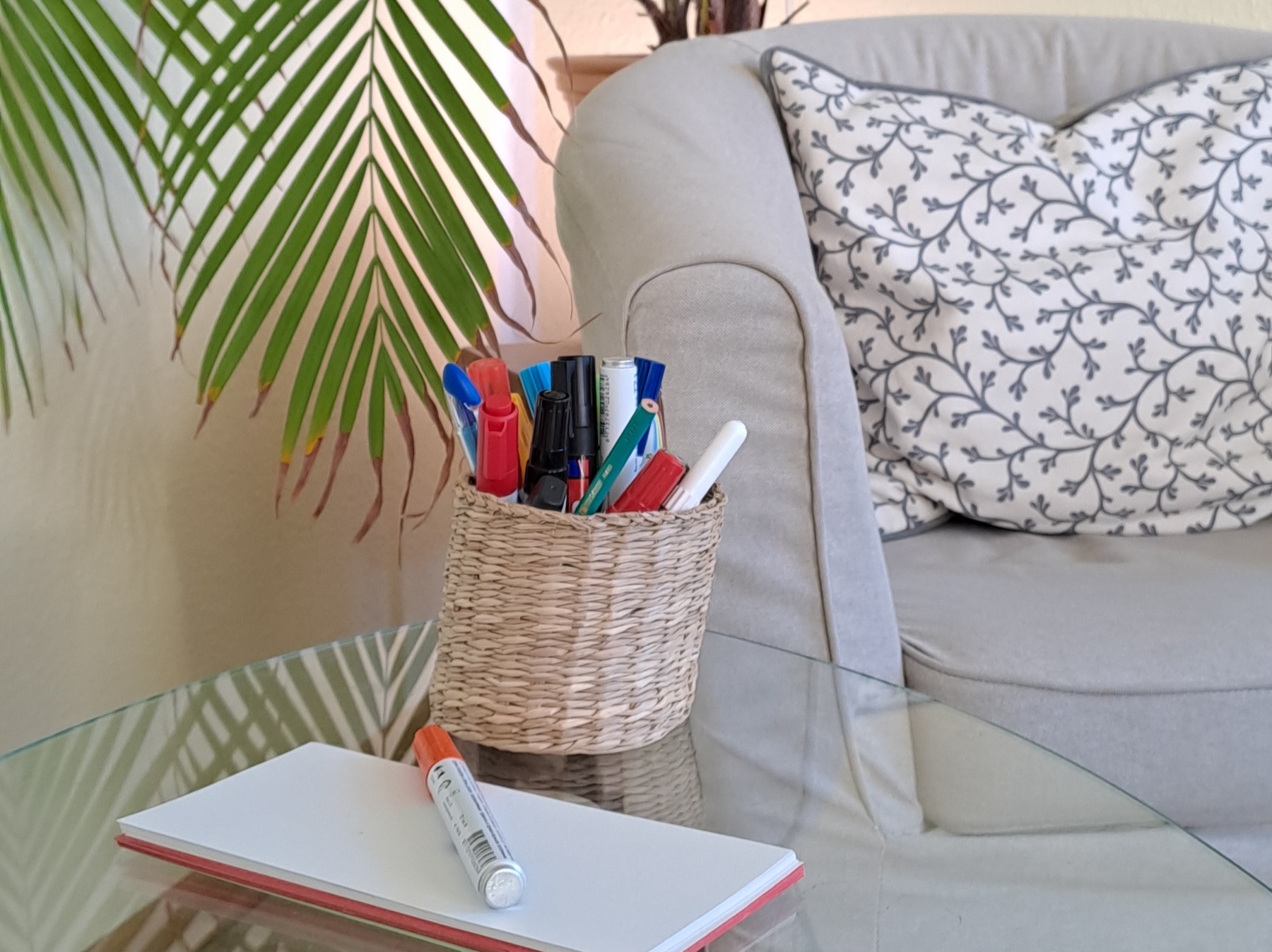 Ein Korb mit verschiedenen Stiften steht auf einem kleinen Tisch. Daneben liegt ein Notizblock mit einem Stift darauf. Im Hintergrund ist ein Teil eines Sofas mit einem Kissen zu sehen. Die Szene wirkt gemütlich und einladend zum Schreiben oder Zeichnen.