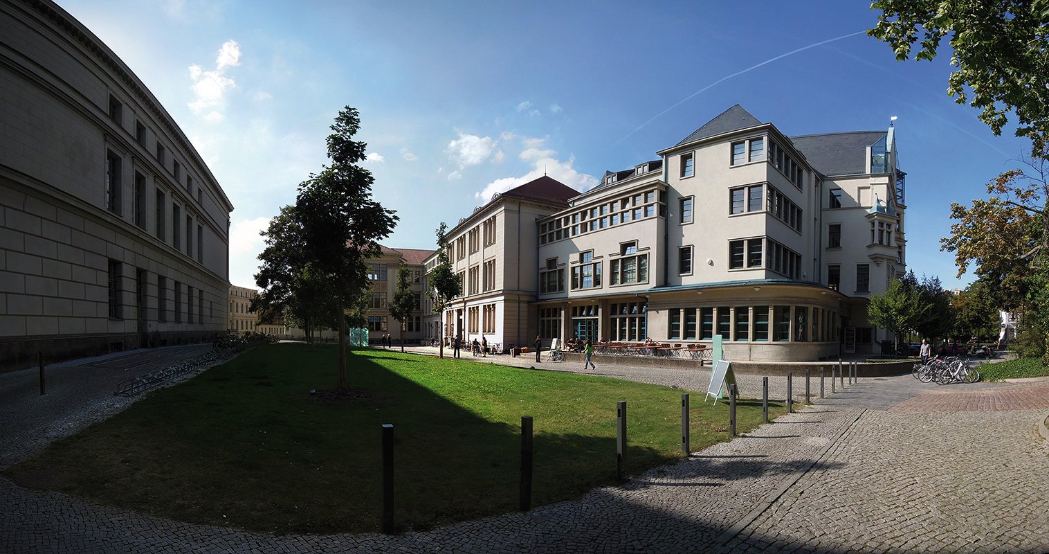 Ein Panoramabild eines Universitätsgeländes mit mehreren Gebäuden, einem gepflegten Rasen und einem gepflasterten Weg. Im Vordergrund befinden sich Bäume und einige Fahrradständer. Der Himmel ist klar mit einigen Wolken.