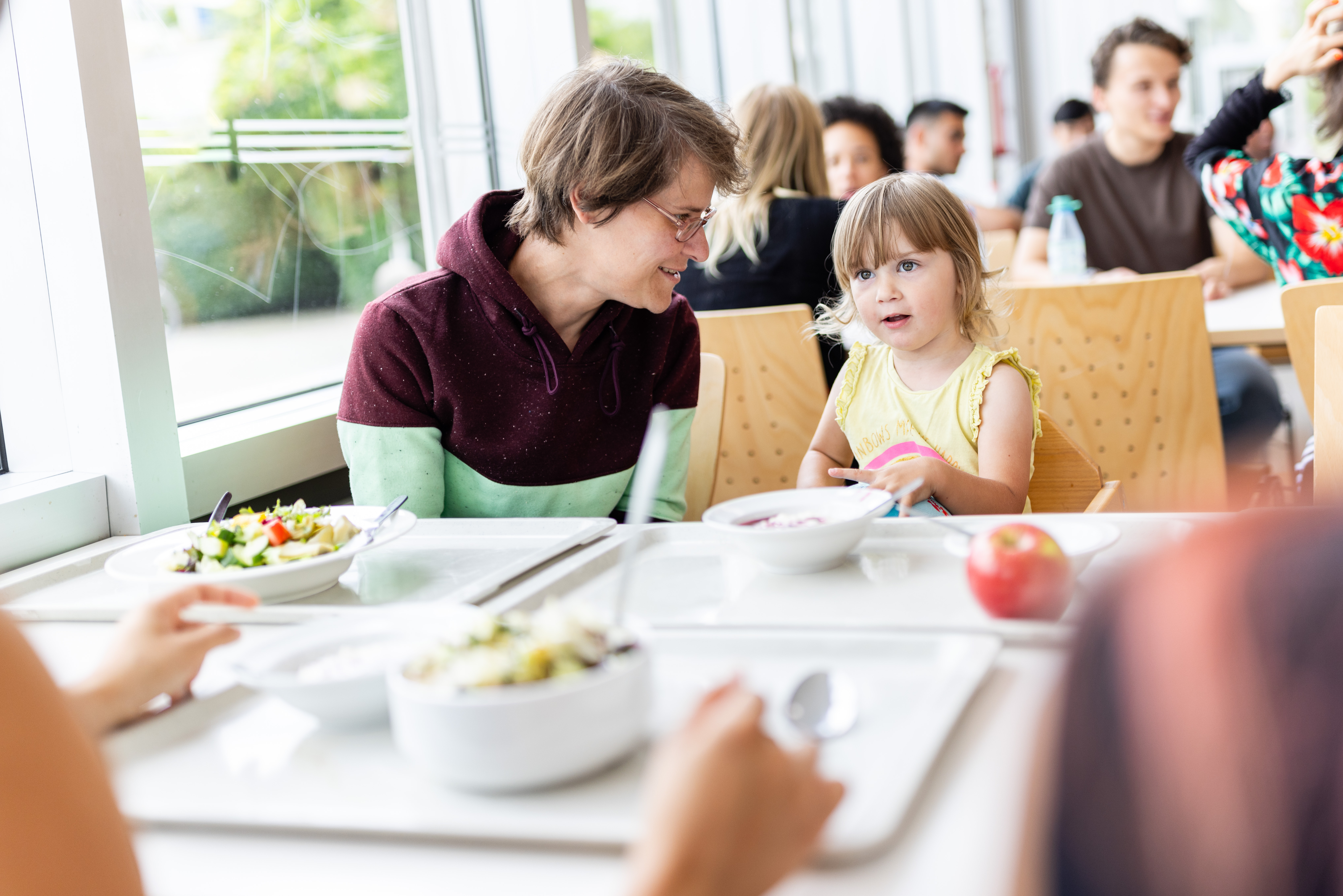 Ein Mann und ein kleines Mädchen sitzen in einem hellen Speiseraum an einem Tisch und essen gemeinsam. Der Mann lächelt das Mädchen an, während sie isst. Auf dem Tisch stehen Schüsseln mit Salat und eine Apfel. Andere Personen sind im Hintergrund zu sehen.