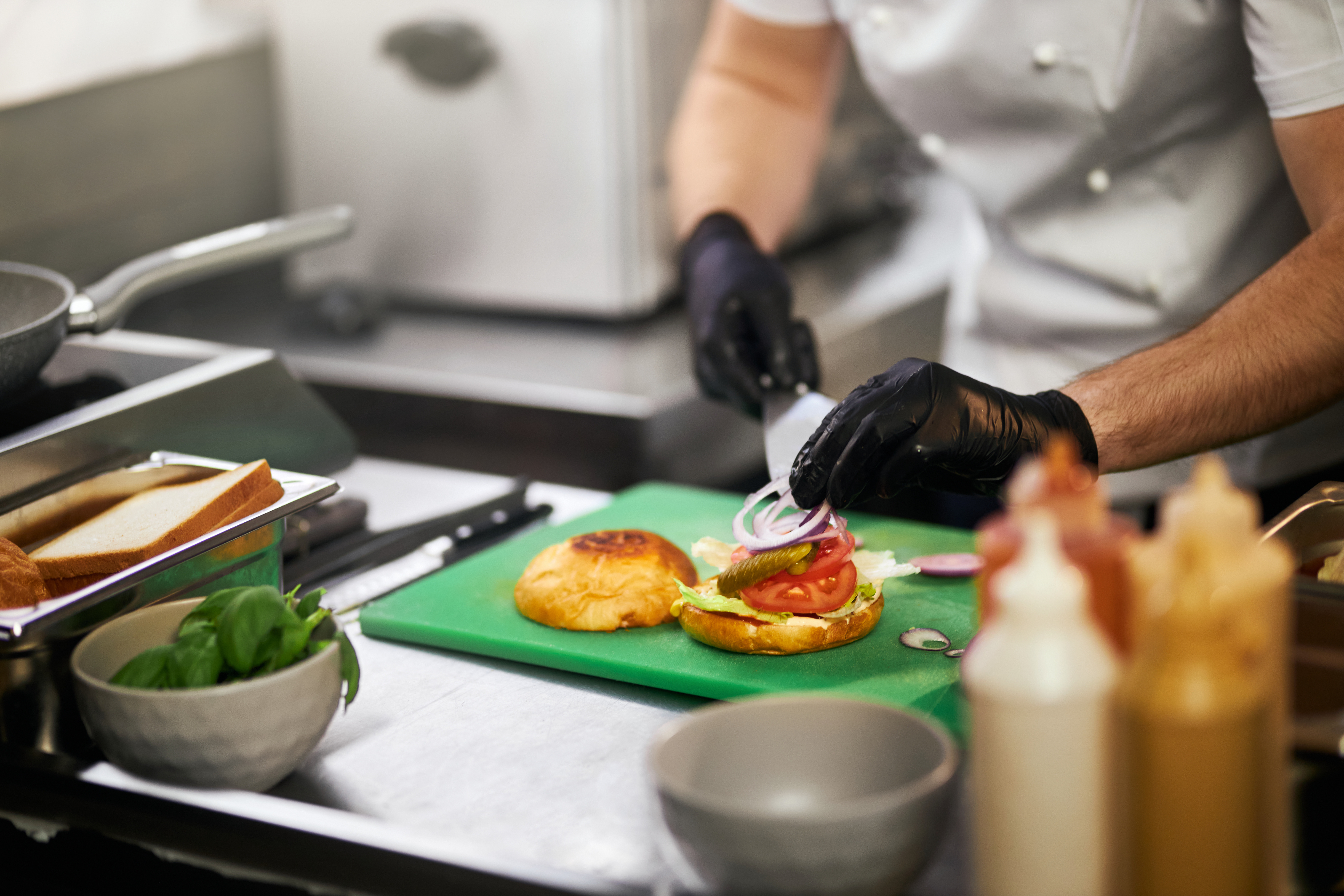 Ein Koch in Schutzhandschuhen bereitet auf einer grünen Schneidunterlage ein belegtes Brötchen mit Tomaten, Salat und einer Sauce zu, während im Hintergrund eine Pfanne und weitere Zutaten zu sehen sind.