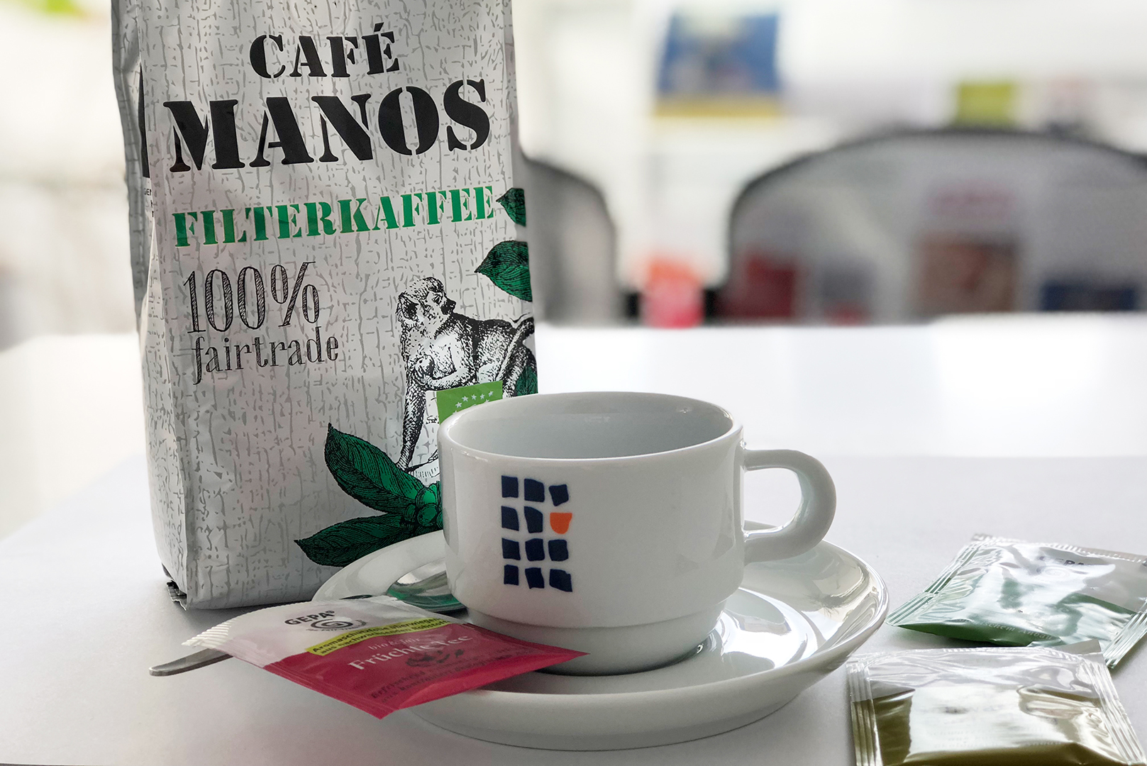 "Ein weißer Kaffeebecher mit einem bunten Logo steht auf einer Untertasse. Daneben liegt ein Beutel mit Café Manos Filterkaffee, der als fairtrade zertifiziert ist. Auf dem Tisch befinden sich außerdem verschiedene Teebeutel und ein Löffel."
