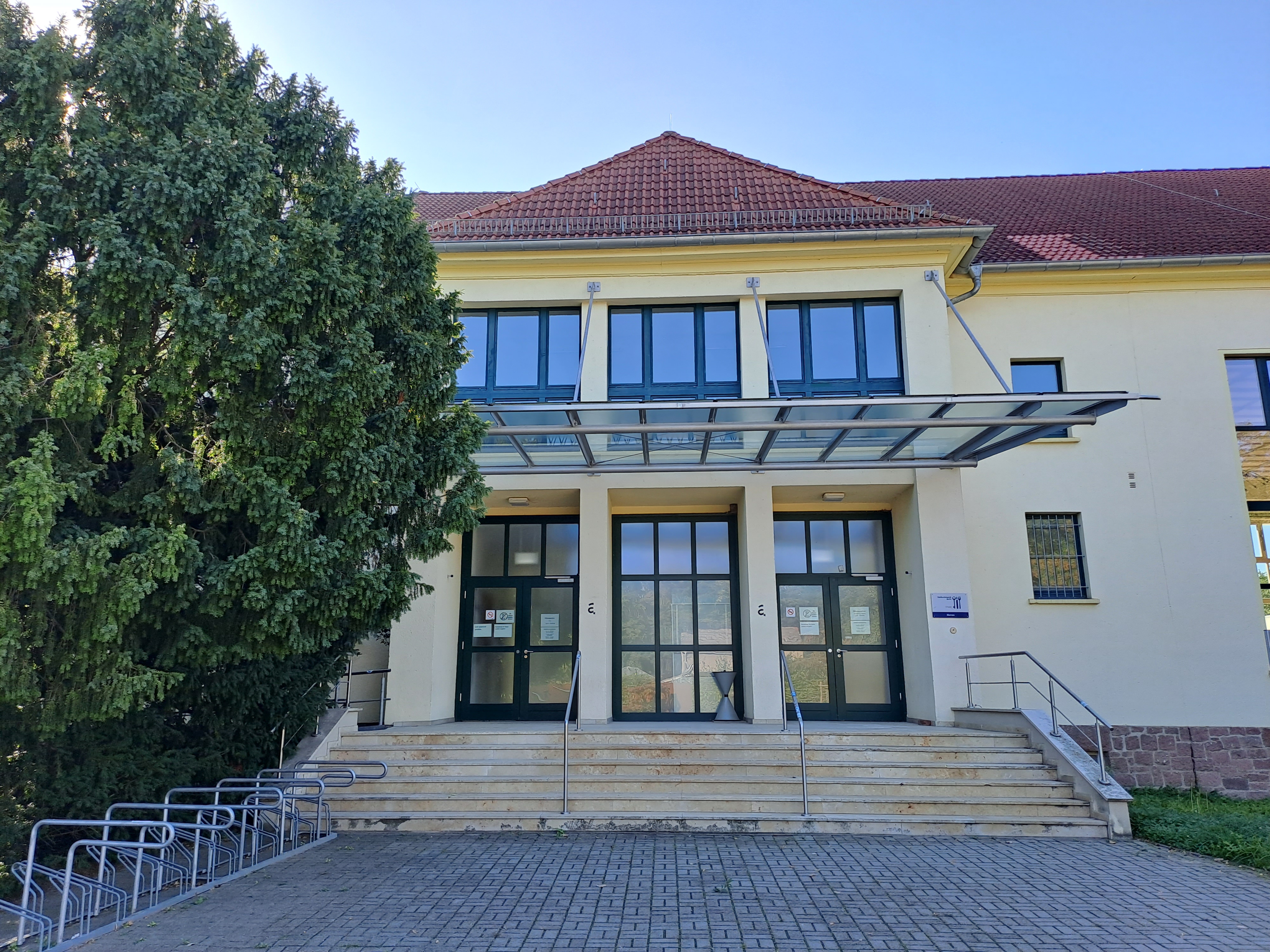 Ein Gebäude mit gelber Fassade, rotem Dach und mehreren großen Fenstern. Der Eingang besteht aus einer doppelten Glastür mit einem überdachten Vordach. Vor dem Gebäude befindet sich ein Baum und eine kleine Treppe mit einem Handlauf. Der Boden ist gepflastert.
