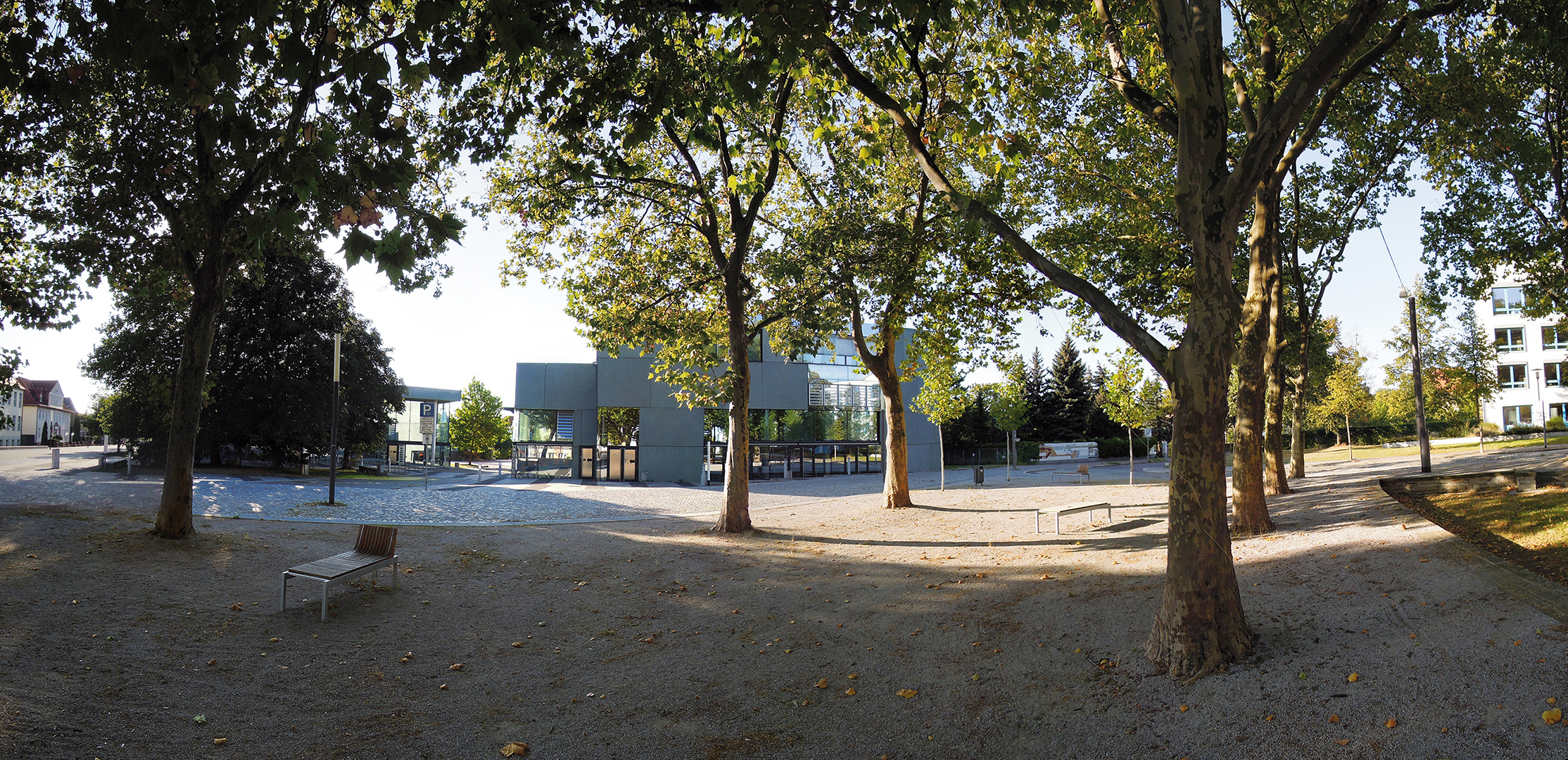 Ein Park mit Bäumen, einer Bank und einem Spielplatz im Hintergrund. Im Hintergrund sind Gebäude zu sehen. Der Boden ist mit Kies bedeckt und es ist ein sonniger Tag.
