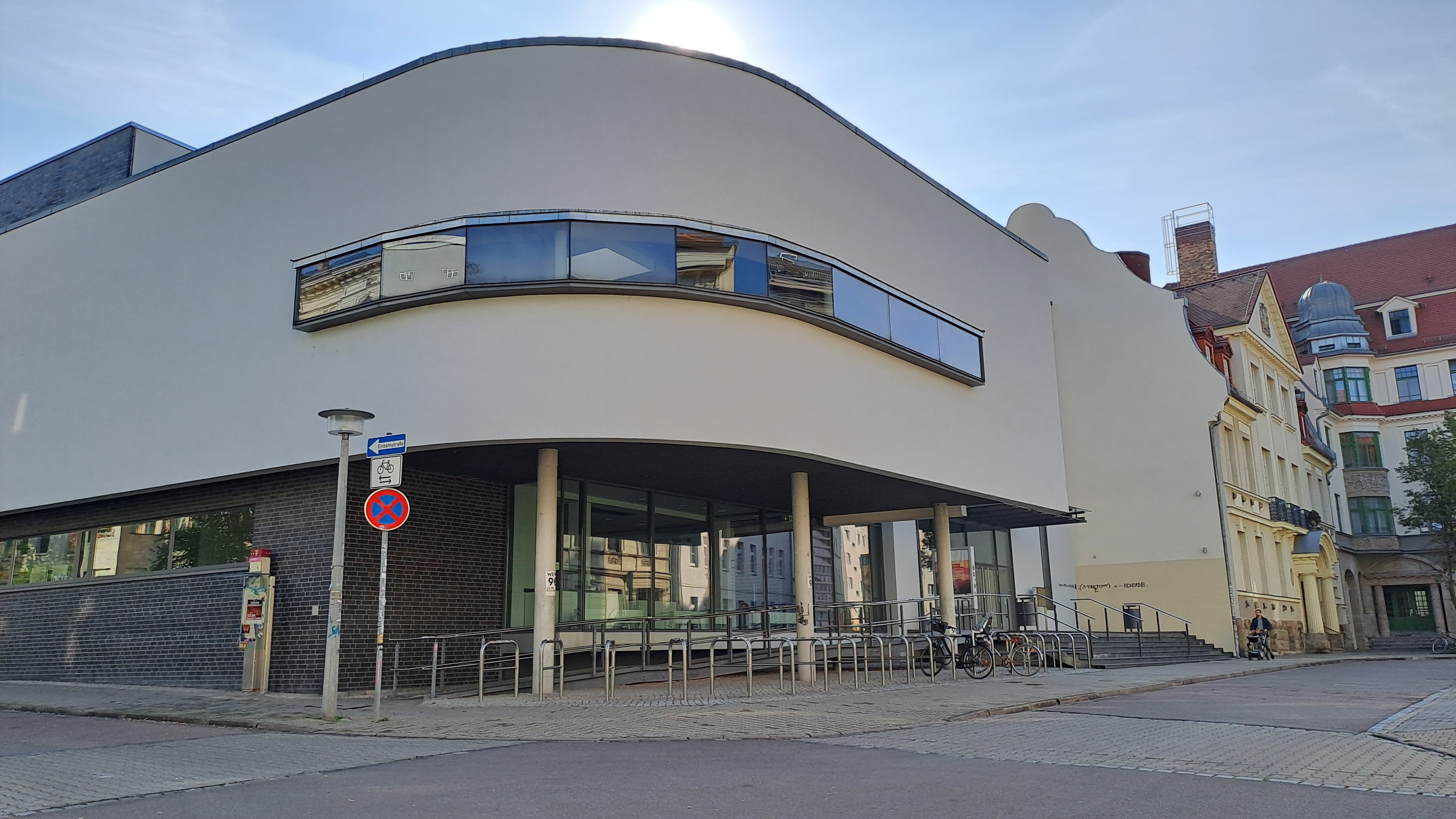 "Modernes Gebäude mit abgerundeten Ecken und großen Fenstern. Vor dem Gebäude befindet sich ein Fahrrad und ein Schild mit der Aufschrift 'Einbahnstraße'. Im Hintergrund sind weitere Gebäude zu sehen."