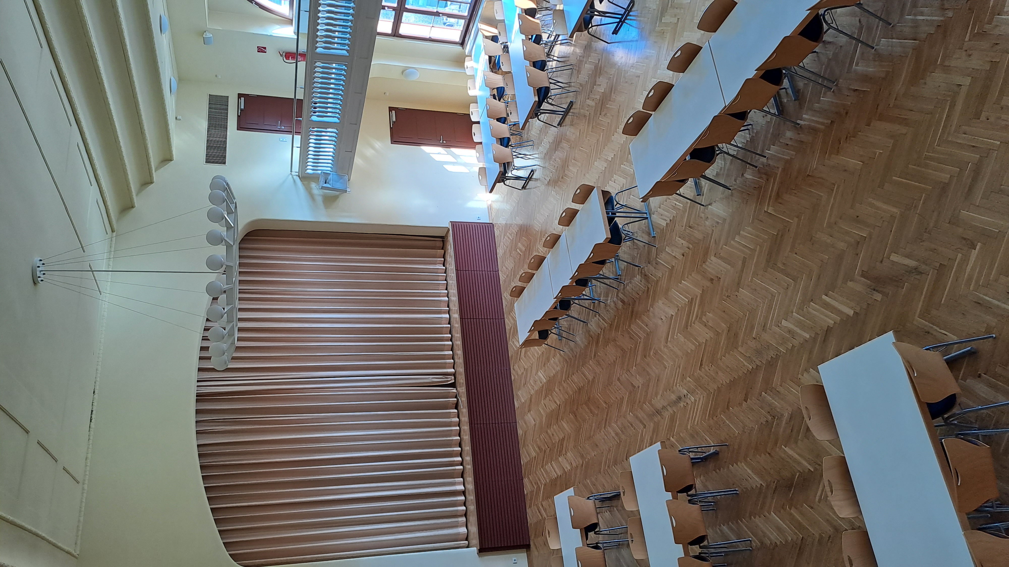 Ein leerer Speisesaal mit mehreren Tischen und Stühlen, die in Reihen angeordnet sind. An einer Wand befindet sich ein großer Vorhang, und im Hintergrund ist eine Galerie mit einem Geländer zu sehen. Die Decke hat eine moderne Beleuchtung. Der Boden ist mit Holzparkett ausgelegt.