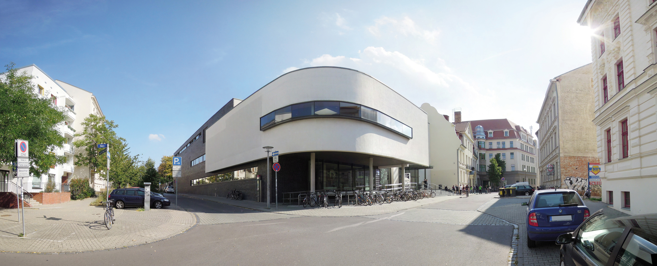 Ein modernes Gebäude mit weißer Fassade steht an einer Straßenecke. Vor dem Gebäude sind Fahrräder abgestellt und ein Auto fährt vorbei. Die umliegenden Gebäude sind aus Backstein und moderner Architektur. Die Szene wirkt ruhig und urban.