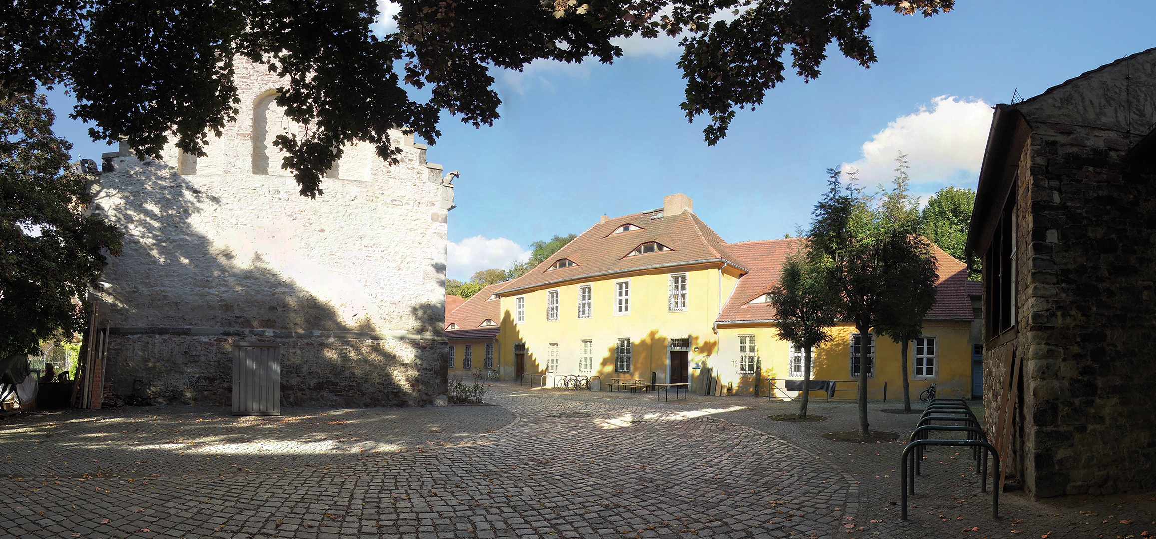 Ein sonniger Platz mit Kopfsteinpflaster, umgeben von historischen Gebäuden. Im Hintergrund steht eine alte, teilweise verfallene Mauer. Ein gelbes Haus mit rotem Dach ist zentral positioniert. Bäume werfen Schatten auf den Platz. Die Atmosphäre wirkt ruhig und malerisch.