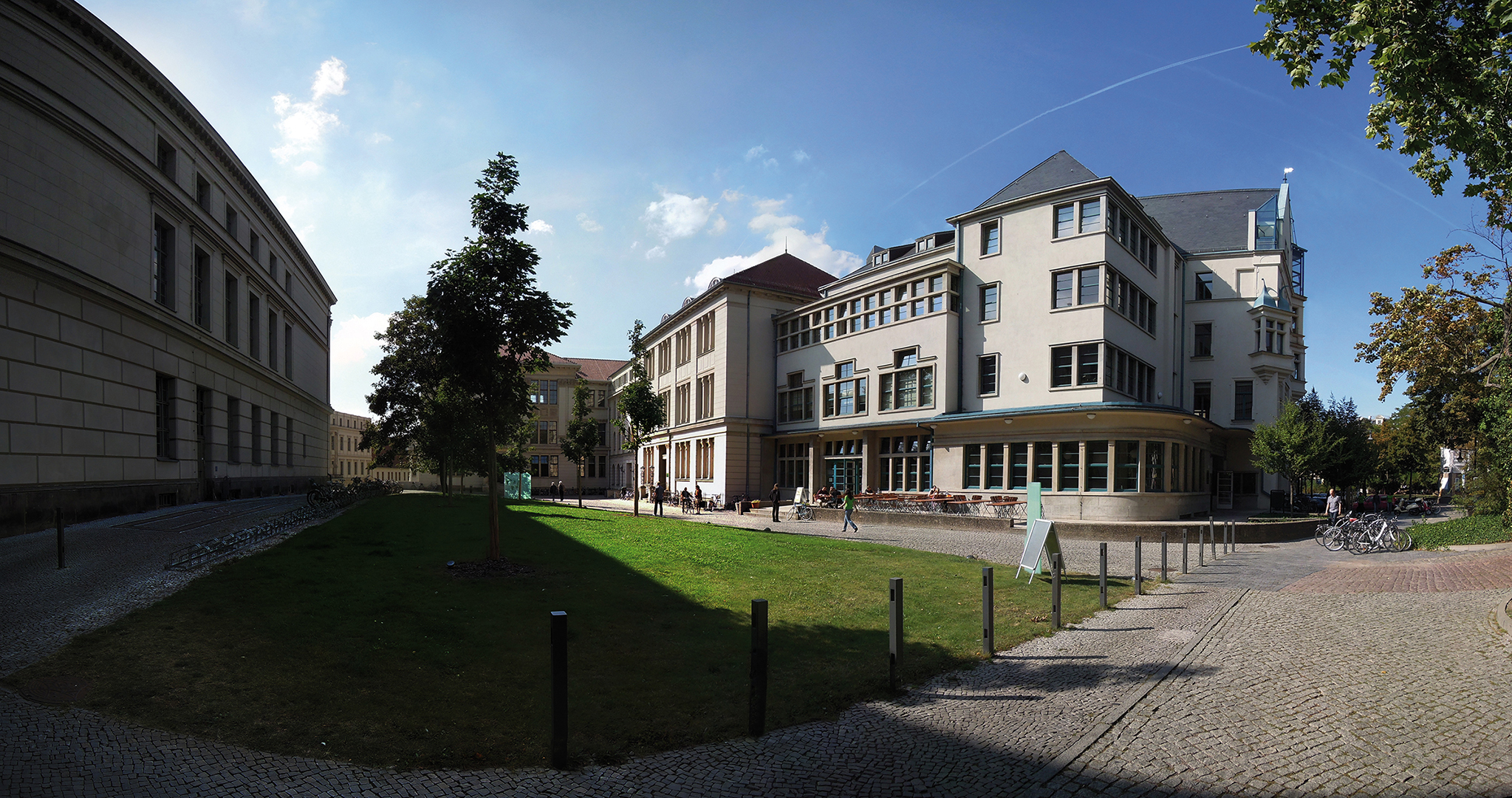 Ein Panoramafoto eines Universitätsgeländes mit mehreren Gebäuden, einem gepflegten Rasen und einem gepflasterten Weg. Im Vordergrund befinden sich Bäume und einige Fahrräder. Der Himmel ist klar mit vereinzelten Wolken. Die Architektur der Gebäude ist modern und funktional.