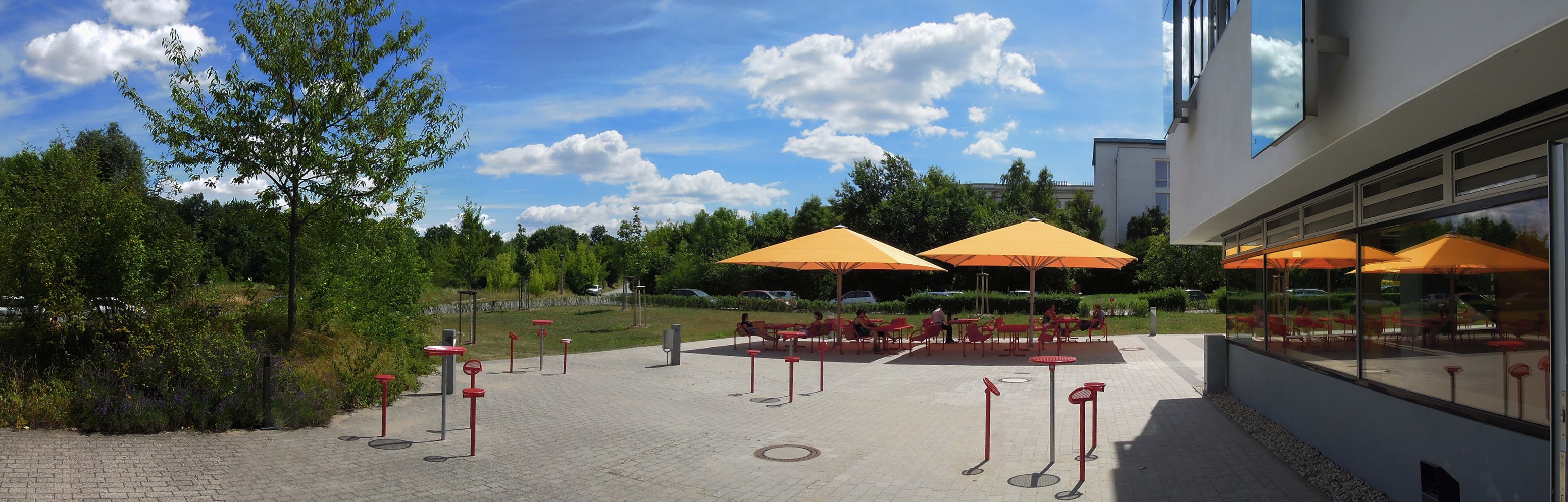 Ein sonniger Außenbereich mit mehreren Tischen und Stühlen unter orangefarbenen Sonnenschirmen. Im Hintergrund sind Bäume und ein modernes Gebäude zu sehen. Der Boden ist gepflastert, und es gibt einige Spielgeräte für Kinder in der Nähe.