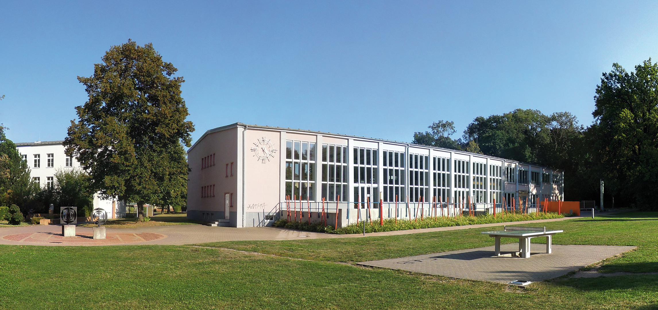 Ein modernes Schulgebäude mit großen Fenstern und einem Uhrturm. Vor dem Gebäude befindet sich ein gepflegter Rasen mit einigen Bäumen und Bänken. Der Himmel ist klar und blau.