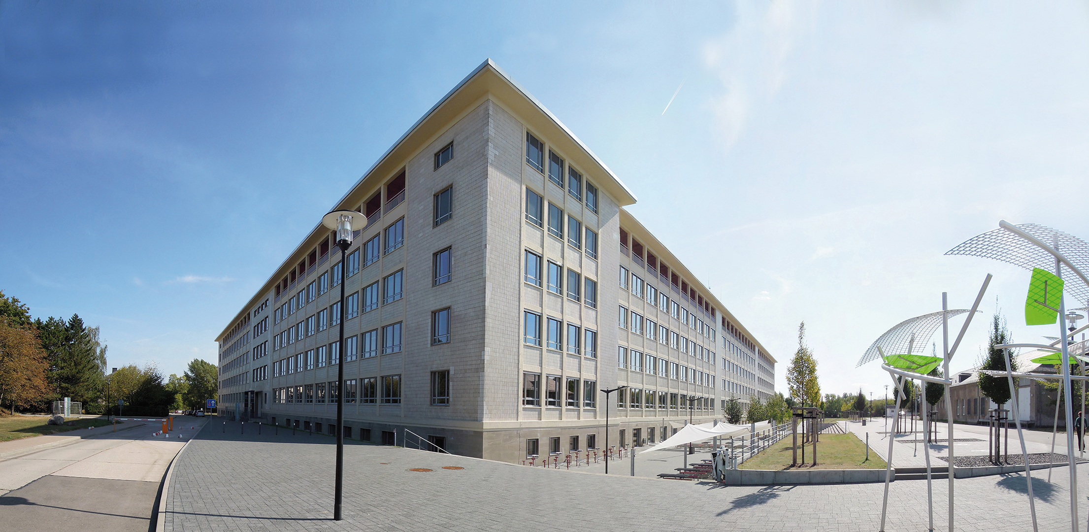 "Ein modernes, mehrstöckiges Gebäude mit vielen Fenstern steht auf einem weitläufigen, gepflasterten Platz. Im Vordergrund befinden sich moderne Sitzgelegenheiten und ein kleiner, grüner Pavillon. Der Himmel ist klar und blau, und es sind einige Bäume im Hintergrund zu sehen."