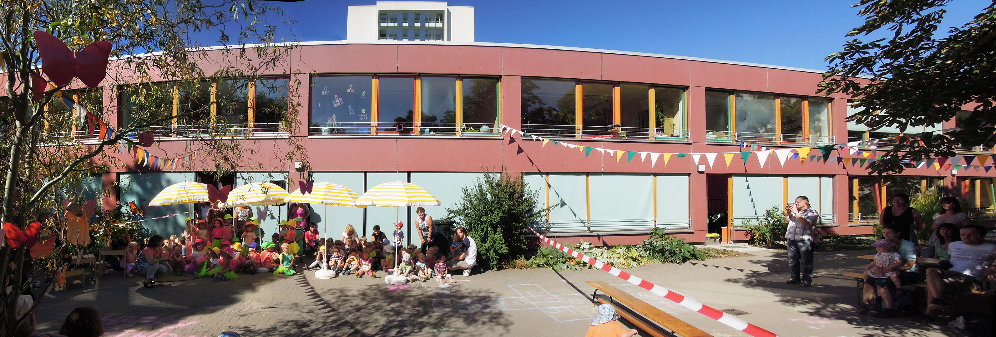 Ein Gebäude mit großer Glasfront und mehreren Fenstern, umgeben von Menschen, die an einem festlichen Event teilnehmen. Bunte Girlanden schmücken die Fassade. Im Vordergrund sind Tische und Stühle aufgestellt, und einige Personen sitzen oder stehen in Gruppen zusammen.
