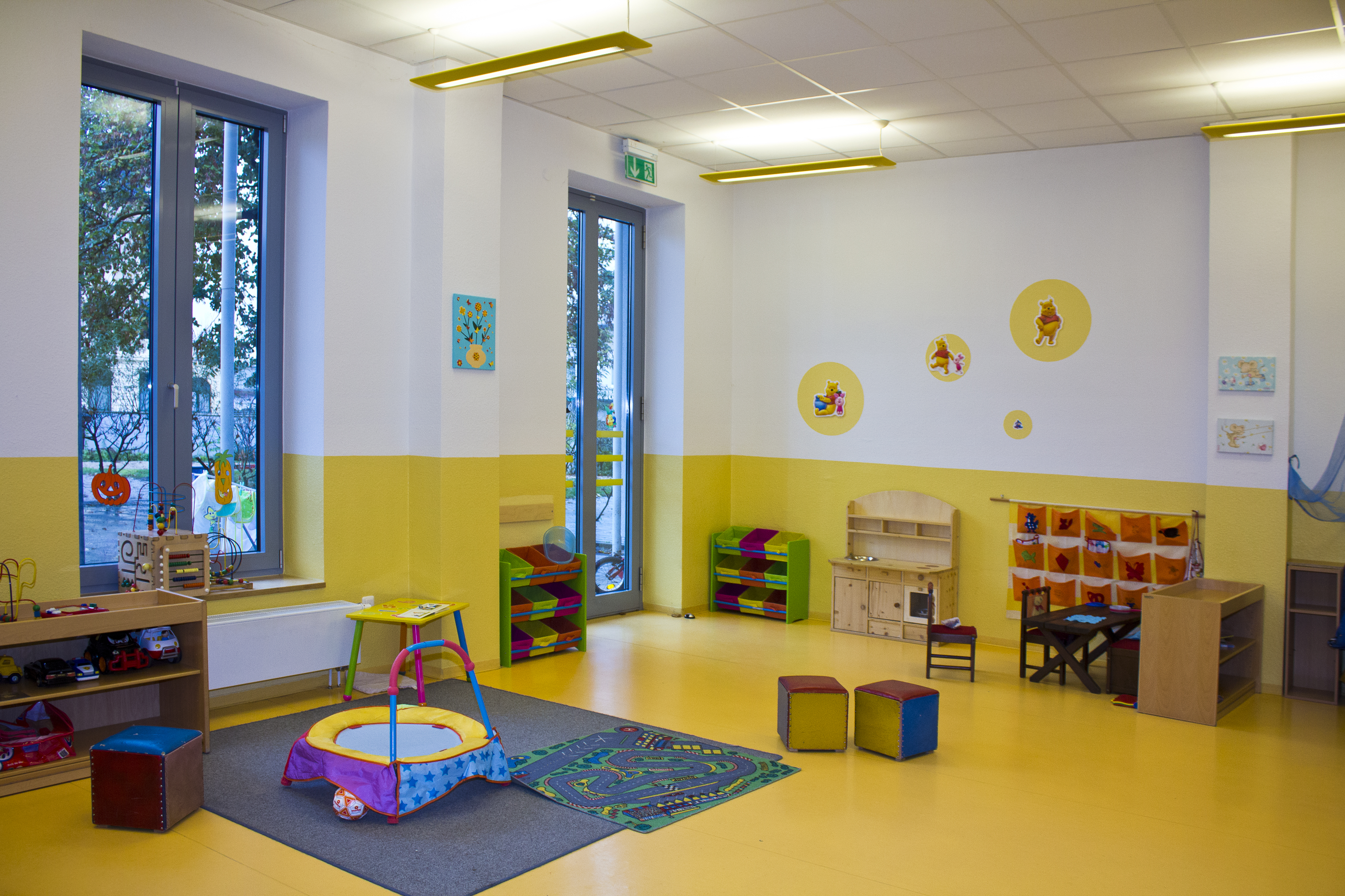 Ein helles, freundliches Kinderzimmer mit gelben Wänden und großen Fenstern. Es gibt Spielzeug, Regale, einen kleinen Tisch mit Stühlen und bunte Bilder an den Wänden. Der Boden ist mit einem Teppich und Spielmatten bedeckt. Das Zimmer ist für Kinder zum Spielen und Lernen eingerichtet.
