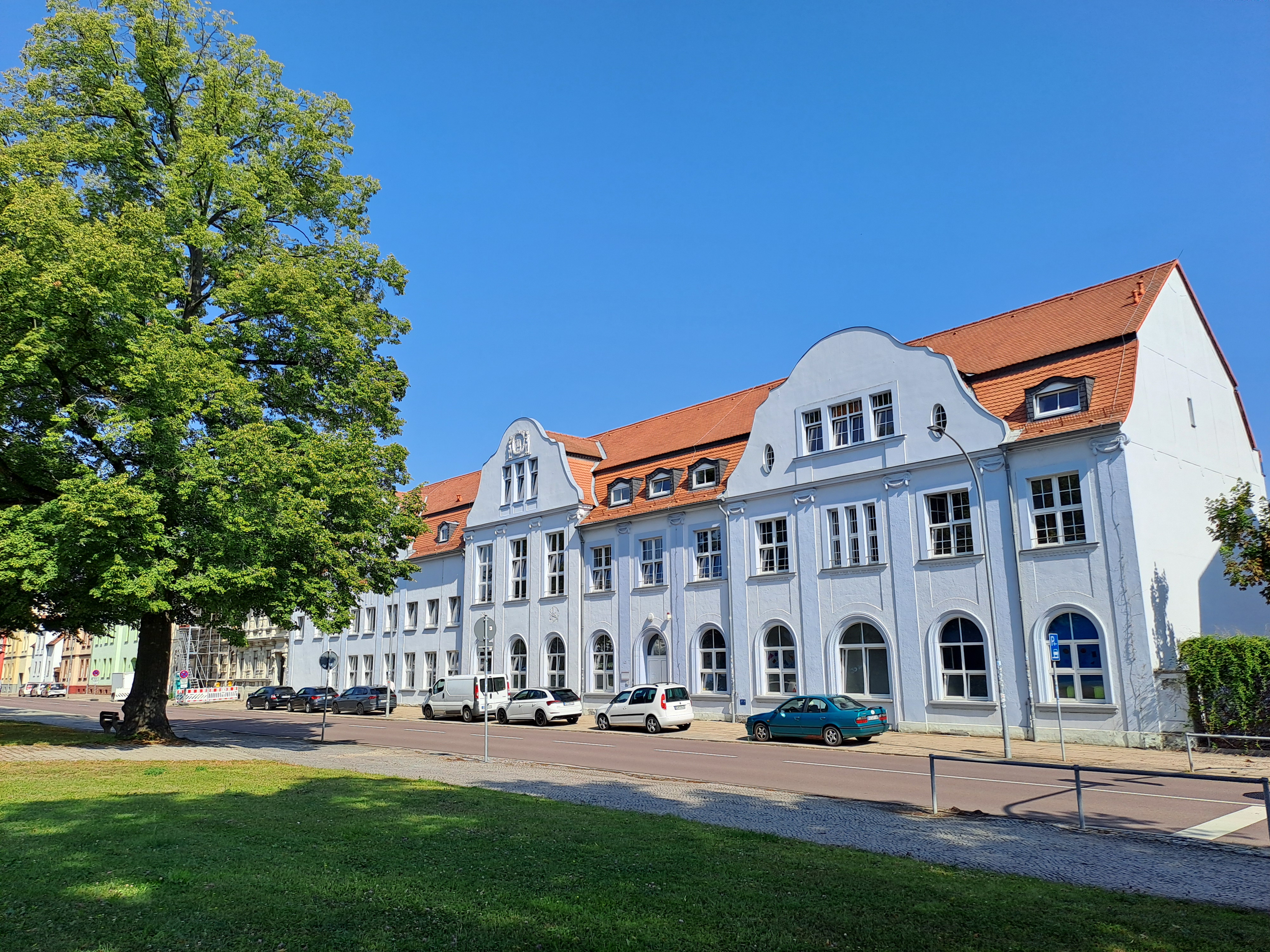 Ein großes, weißes Gebäude mit rotem Dach und vielen Fenstern. Vor dem Gebäude stehen mehrere Autos auf einem Parkplatz. Links im Bild befindet sich ein großer Baum. Der Himmel ist klar und blau, und es gibt einen grünen Rasen im Vordergrund.