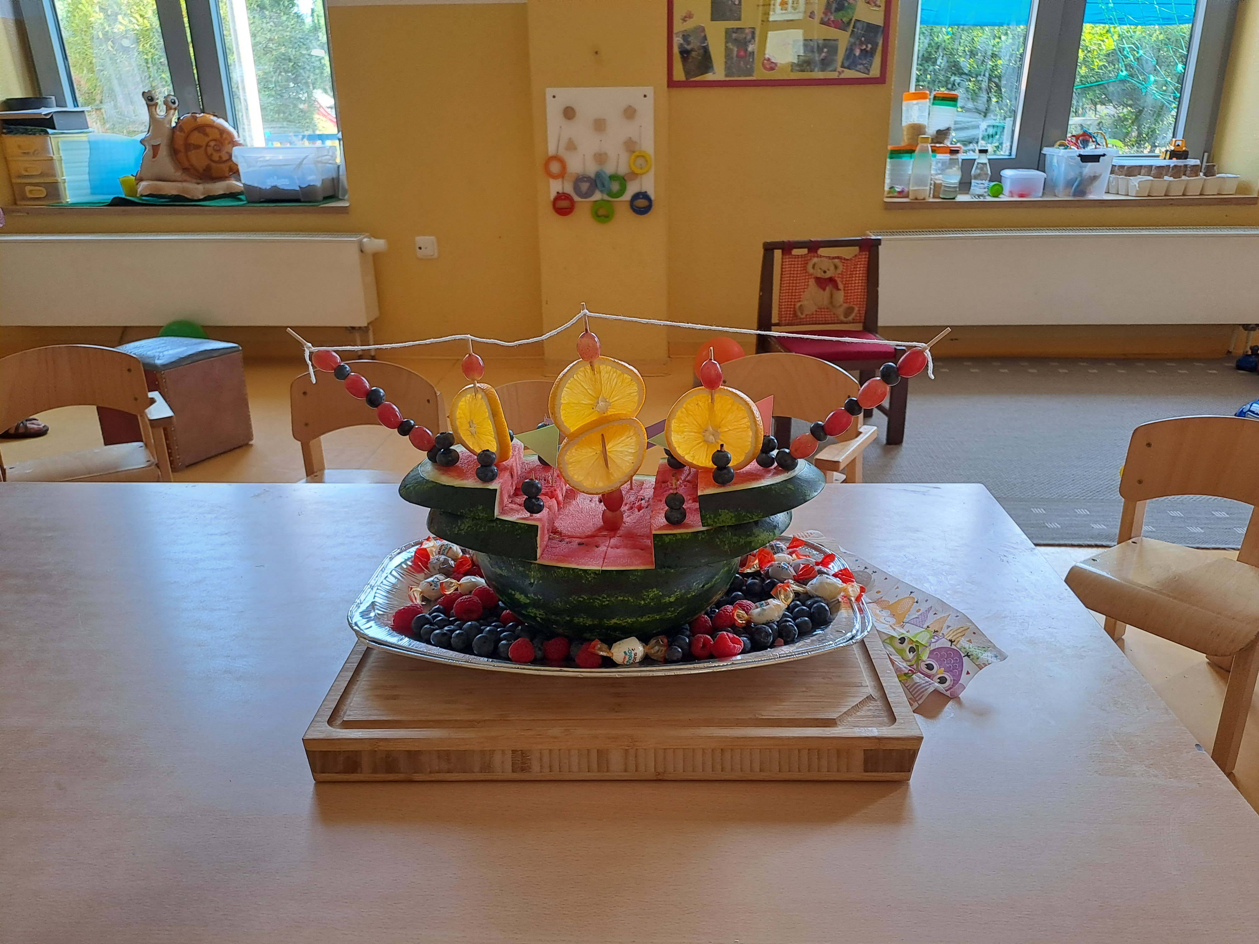 Ein kreativer Obstteller in Form eines Drachen, der mit verschiedenen Früchten wie Trauben, Erdbeeren und Melone dekoriert ist. Der Drache hat bunte Flügel und einen Schwanz, die aus Obst und kleinen Flaggen bestehen. Der Teller steht auf einem Holztisch in einem hellen Raum.