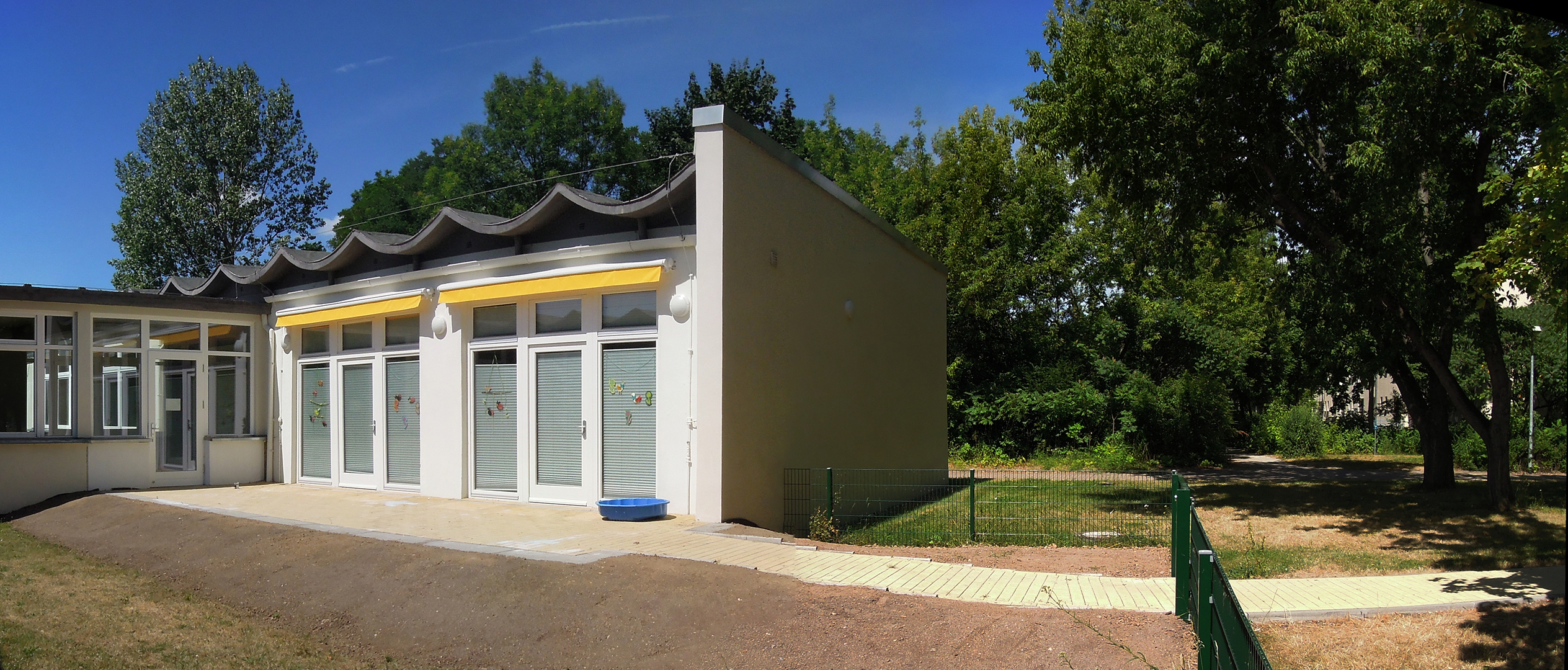 Ein kleines, modernes Gebäude mit gelber Fassade und weißem Dach. Es hat drei große Glastüren und befindet sich in einer grünen, baumbestandenen Umgebung. Der Boden ist gepflastert und es gibt einen kleinen Weg daneben.