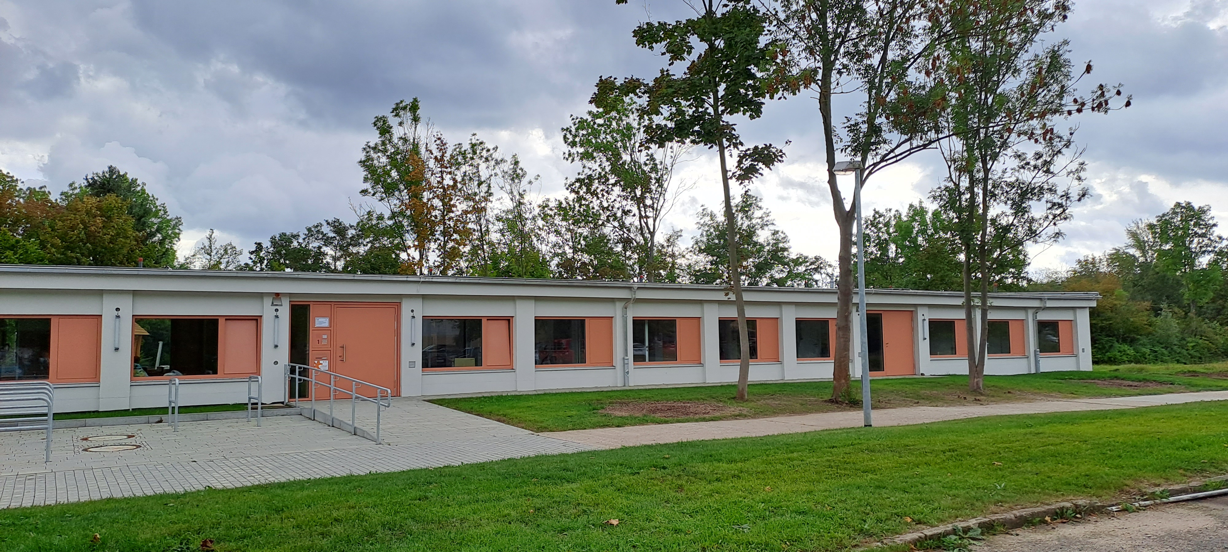 Ein langes, niedriges Gebäude mit orangefarbenen und weißen Wänden, mehreren Fenstern und einer Rampe am Eingang. Das Gebäude steht auf einem grünen Rasen mit einigen Bäumen im Hintergrund. Der Himmel ist bewölkt.