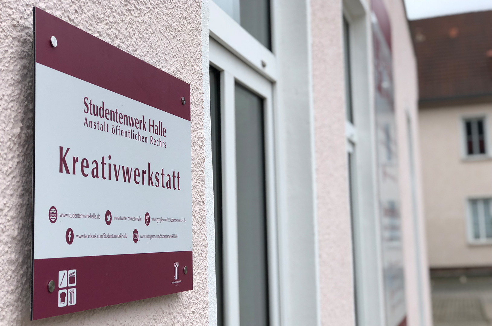 "Ein Schild an einer Wand mit der Aufschrift 'Kreativwerkstatt'. Darunter sind Kontaktdaten und Symbole für Telefon, E-Mail und eine Website. Das Schild gehört zum Studentenwerk Halle, Anstalt öffentlichen Rechts."