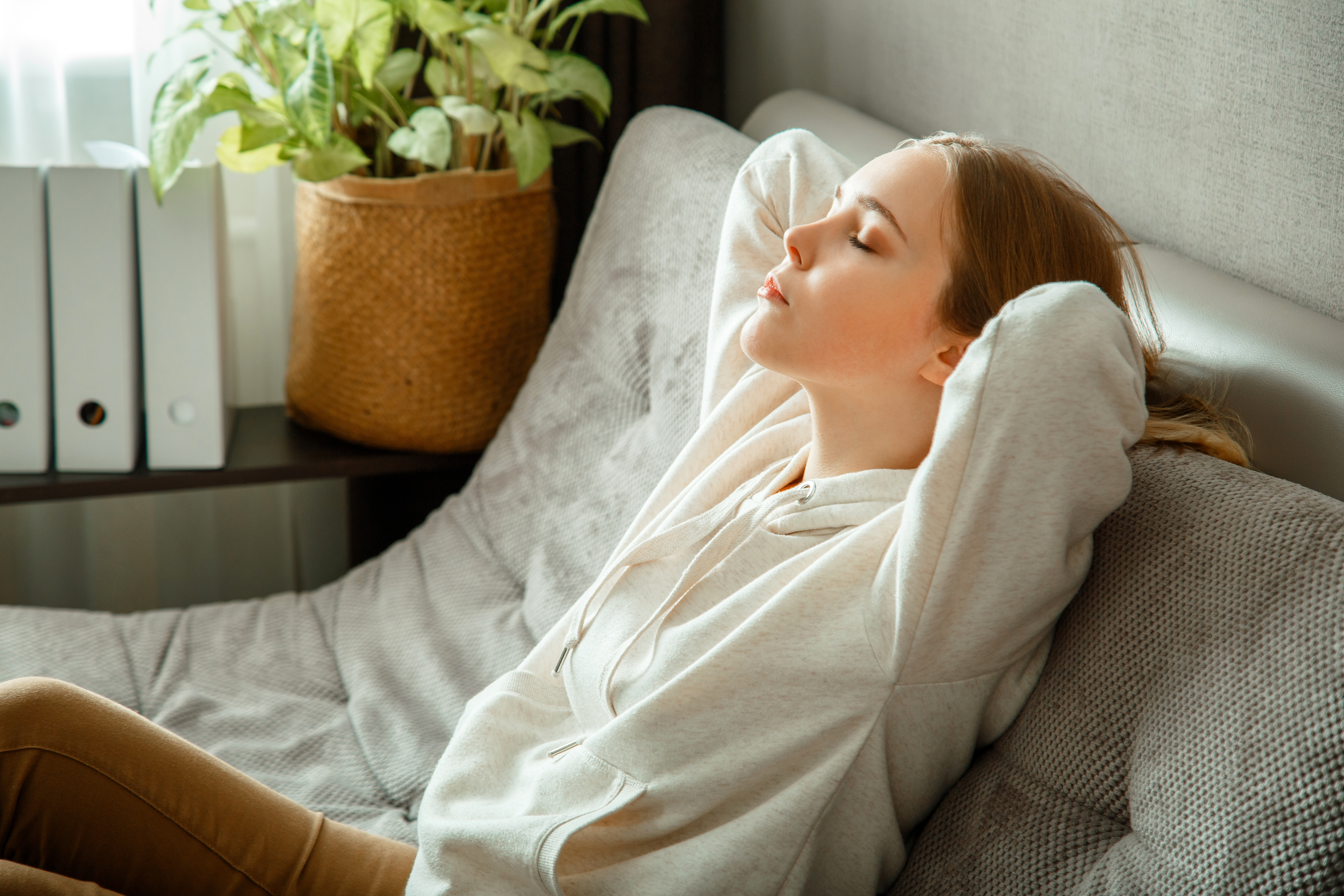 Eine Frau liegt entspannt auf einem Sofa, die Augen geschlossen, und scheint zu meditieren oder sich auszuruhen. Sie trägt einen hellen Hoodie und hat die Hände hinter dem Kopf verschränkt. Der Raum wirkt ruhig und gemütlich.