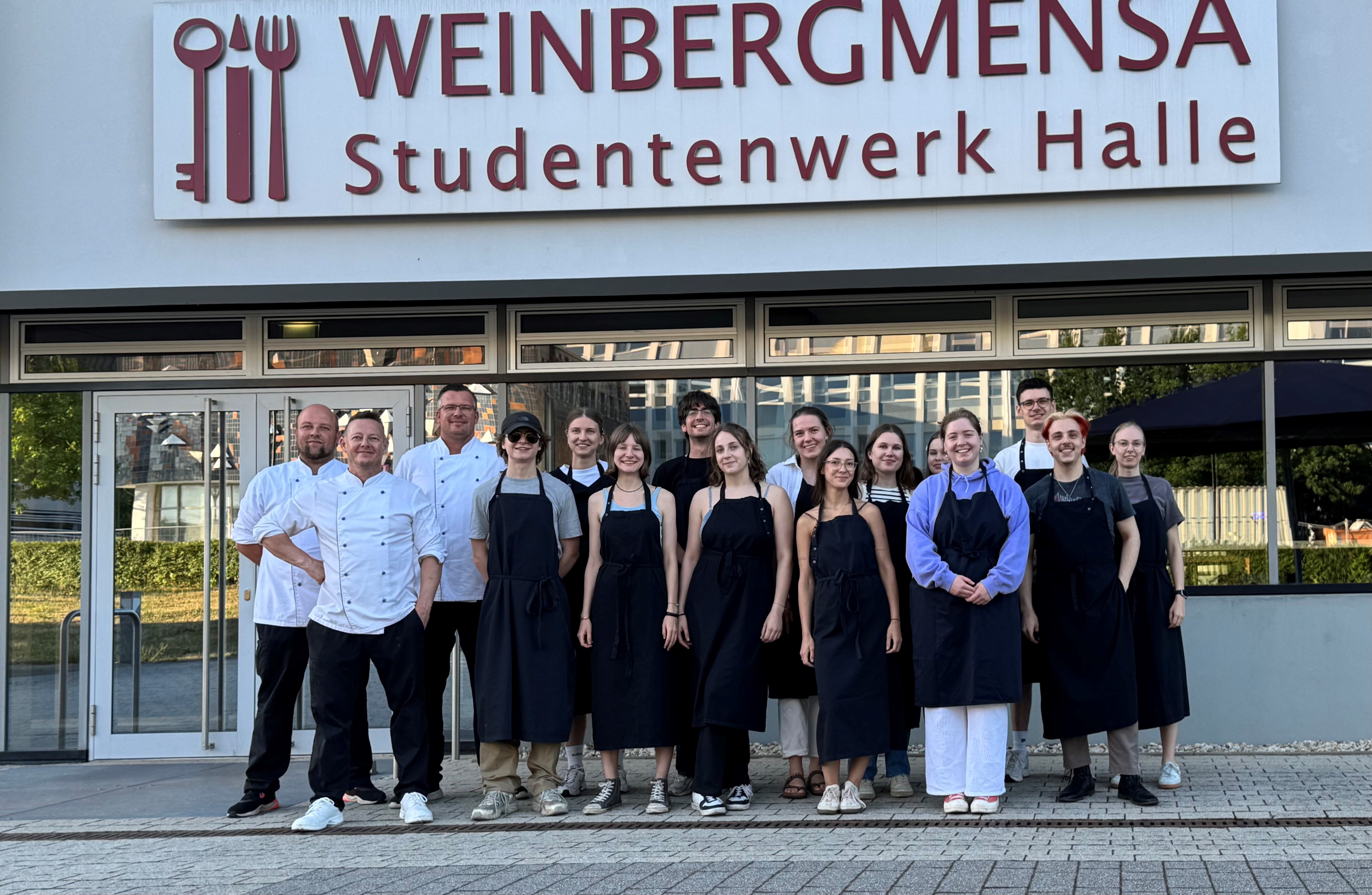 Ein Gruppenfoto vor dem Eingang der "Weinbergmensa Studentenwerk Halle". Die Gruppe besteht aus Mitarbeitern in Koch- und Servicekleidung. Sie stehen vor dem Gebäude und lächeln in die Kamera.