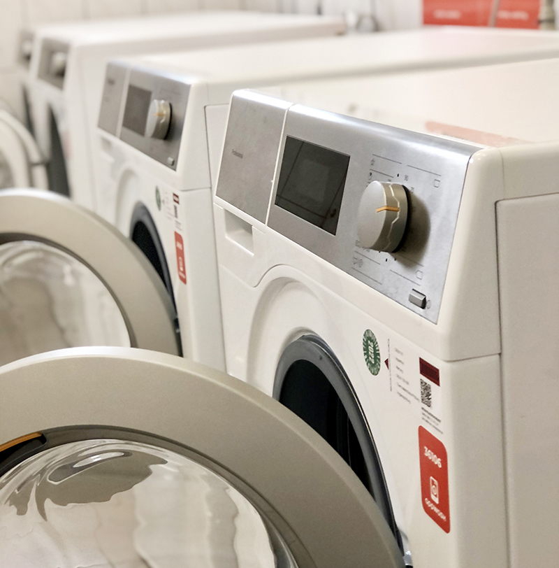"Mehrere weiße Waschmaschinen in einer Reihe, mit digitalen Bedienfeldern und Drehknöpfen. Die Maschinen haben eine Frontlader-Tür und sind in einem gut beleuchteten Raum aufgestellt. Auf den Maschinen sind verschiedene Bedienelemente und Symbole zu sehen."