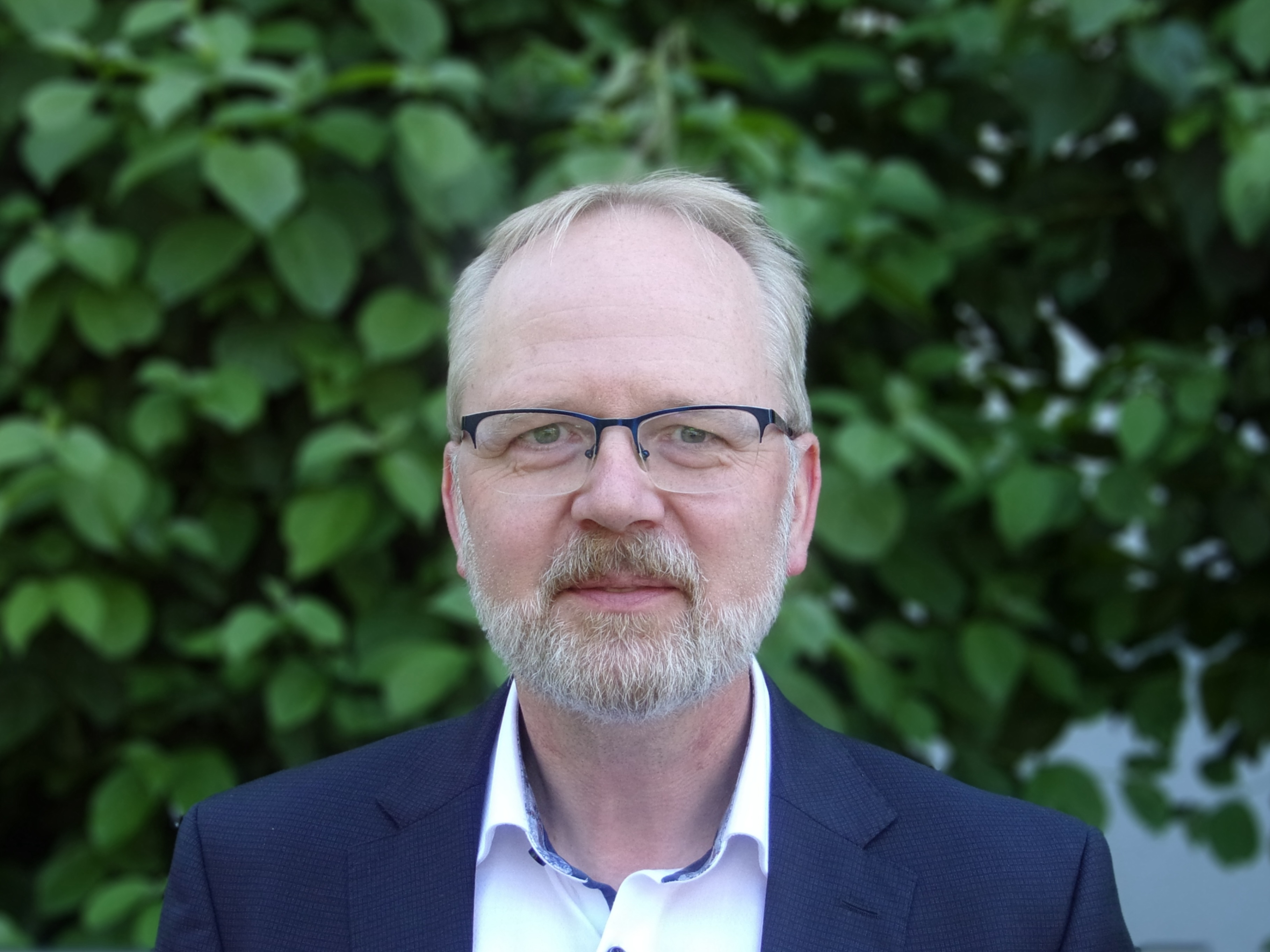 Ein älterer Mann mit grauen Haaren, Brille und grauem Bart trägt ein blaues Sakko und ein weißes Hemd. Er steht vor einem grünen, belaubten Hintergrund.