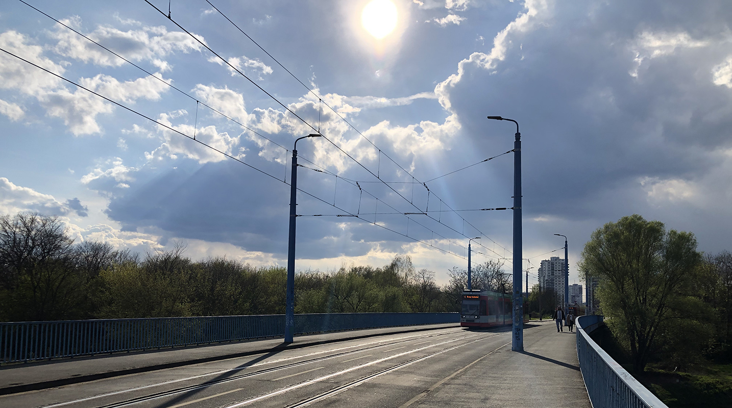 Eine Brücke mit Straßenbahnschienen und Oberleitungen unter einem bewölkten Himmel mit Sonnenlicht, das durch die Wolken scheint. Im Hintergrund sind Bäume und Gebäude zu sehen. Die Brücke ist leer, mit Ausnahme eines fernen Straßenbahnwagens.